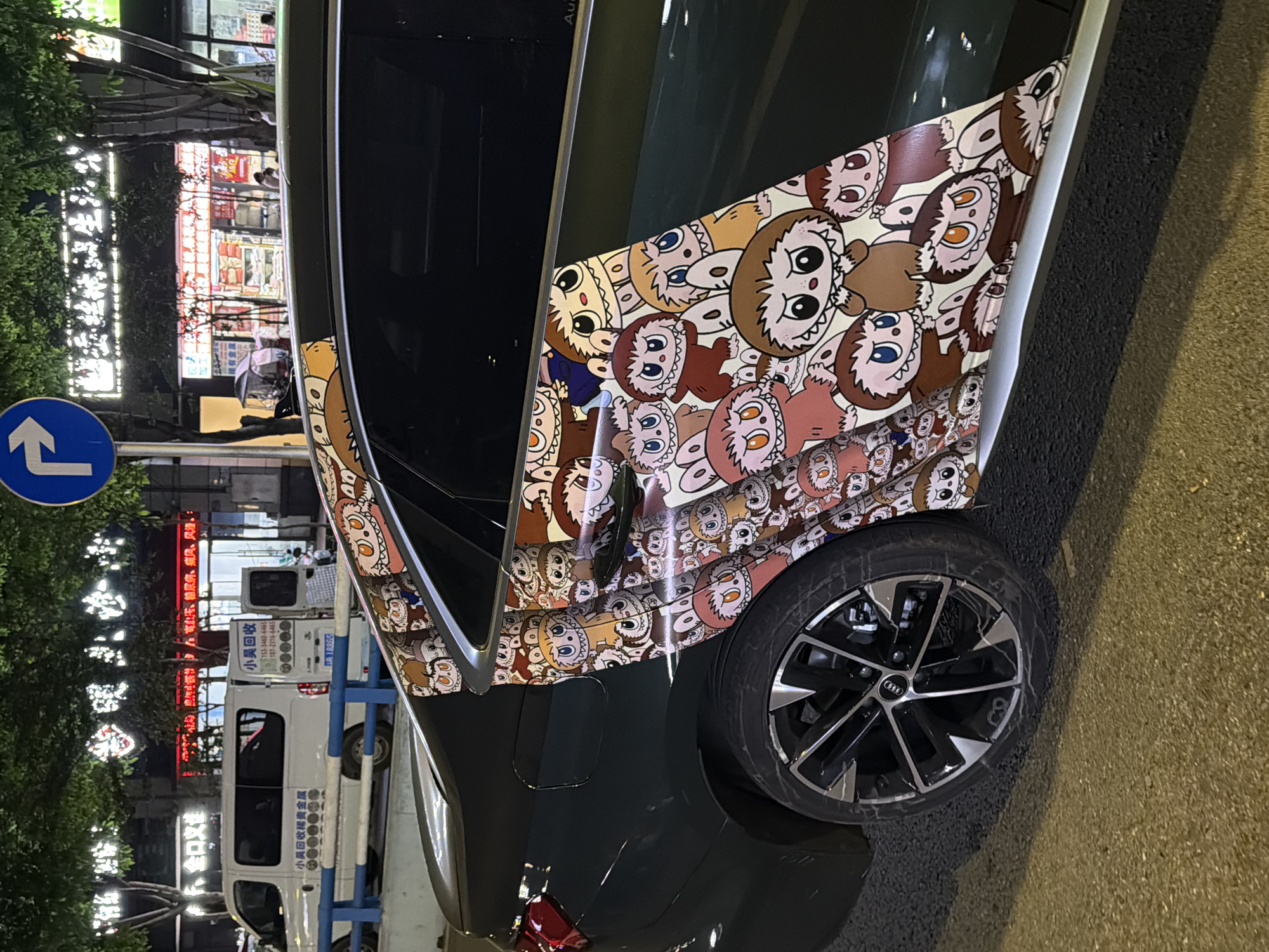 Here's a detailed description of the image:
**General Scene:**
The image shows the rear passenger side of a dark-colored car parked on an asphalt street. The car's exterior is uniquely decorated with a cartoonish pattern of faces. The background shows an urban street scene at night, presumably in Chongqing High-tech Zone, China.
**Specific Details:**
* **Car:** The car appears to be an Audi, identifiable by the wheel design. The surface of the car from the rear passenger door to the rear wheel well is covered with a cartoon graphic.
* **Background:** The background includes a white van that has Chinese characters on it that appear to reference metal recycling. There are trees and what look like shopfronts or commercial buildings. Red neon signs are visible. A blue traffic sign is also visible in the background.
* **Environment:** The scene suggests an urban area with street lighting. The presence of the white van and other commercial signs suggests a potentially busy or mixed-use area. The photograph was most likely taken at night.