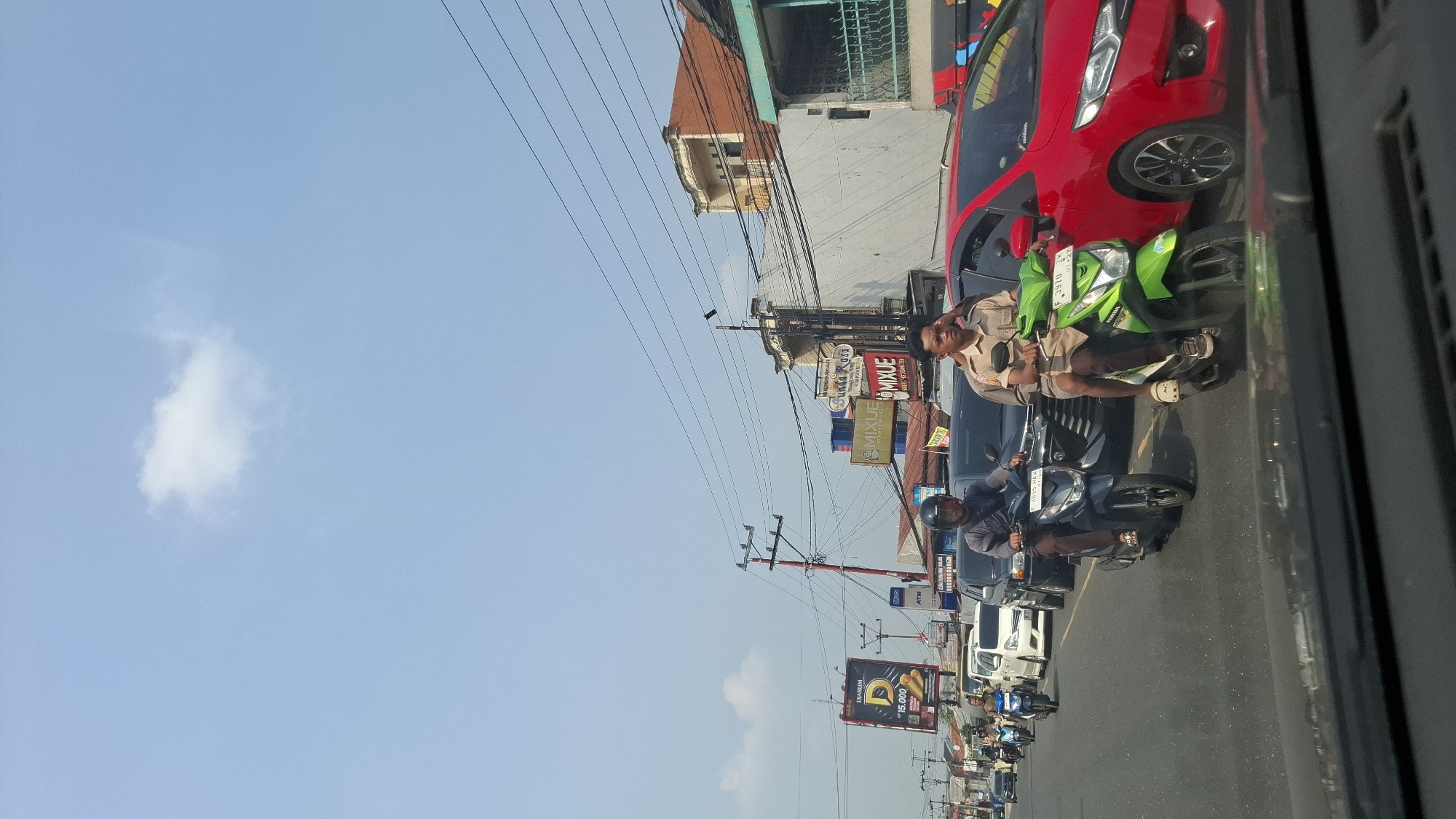 The image depicts a street scene in Cikaroya, Indonesia, on a bright, sunny day with a clear blue sky and a few scattered clouds. The viewpoint is from inside a vehicle, looking forward. The road is busy with traffic, primarily motorbikes and cars.
In the foreground, two motorbikes are prominent. The one on the left, a dark colored scooter, is ridden by a man wearing a dark helmet and a grey shirt, with brown pants. He is wearing sandals. The motorbike on the right is a bright green scooter, ridden by a young man in a light brown uniform, possibly a school uniform or employee attire, with shorts. He is also wearing sandals. Both riders are in motion, navigating through the traffic.
Behind the motorbikes, several cars are visible, including a dark SUV and a red hatchback on the right side of the frame. Further down the road, more vehicles and motorbikes are lined up. The left side of the street features buildings and advertisements. A large billboard displays "DJARUM D 15.000" with an image of cigarettes. Other signs include "ATM" and "MIXUE." The buildings are a mix of styles, with some featuring red-tiled roofs. Utility poles with numerous wires crisscross the scene, indicating the urban environment. The overall impression is of a bustling street during daylight hours.
