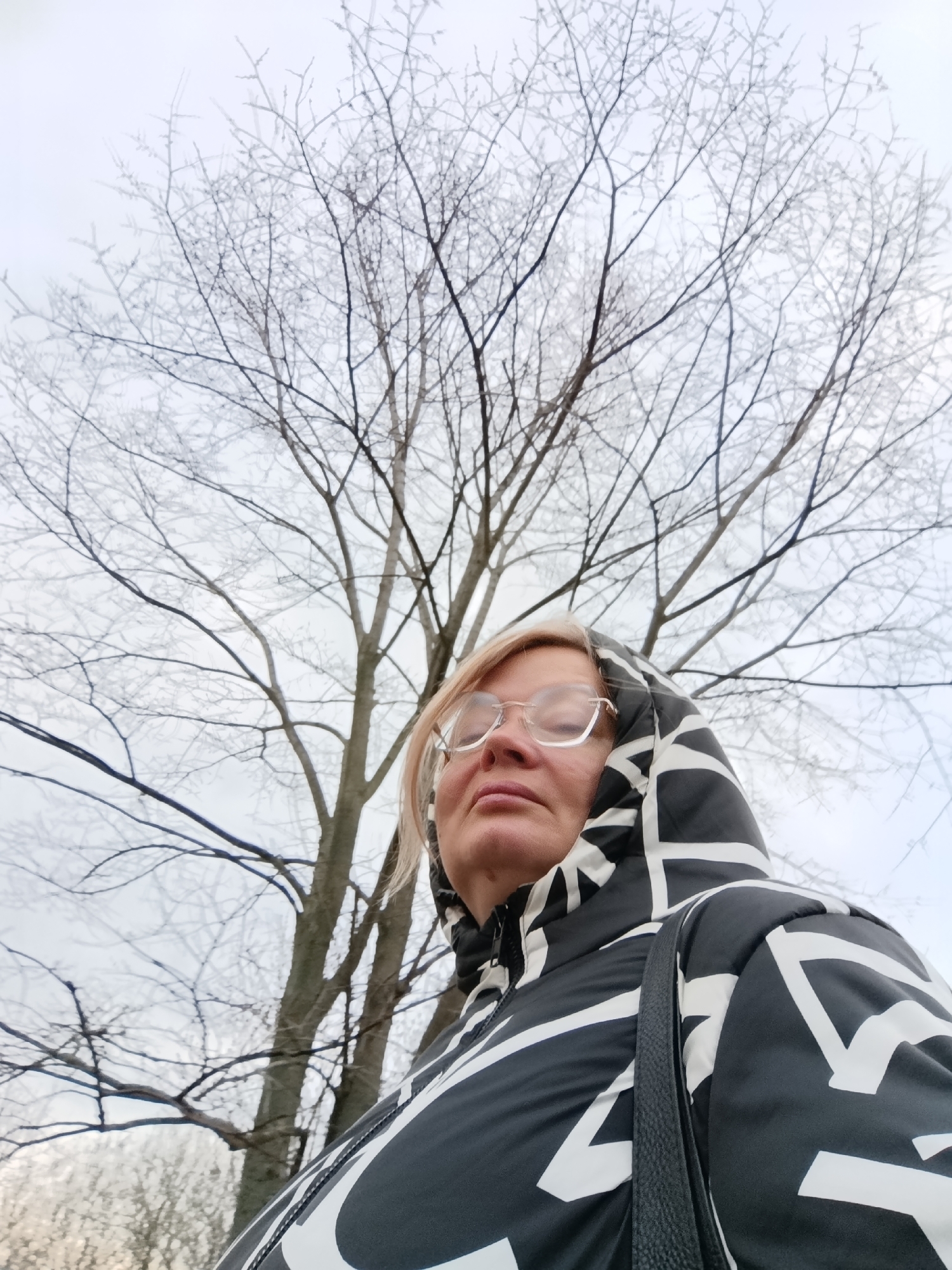 A woman is captured in a low-angle shot, looking upwards and slightly to her left, positioned in the lower right corner of the frame. She has blonde hair and is wearing clear-framed glasses. Her eyes appear narrowed or partially closed as she gazes towards the sky or the tree canopy above.
She is wearing a hooded winter jacket with a striking black and white geometric pattern, where white lines and shapes contrast against a black background. A black shoulder bag strap is visible over her right shoulder.
The background is dominated by a dense network of bare tree branches, reaching towards the overcast, light grey sky. These branches are visibly coated in a delicate layer of rime ice or frost, giving them a distinct white, almost shimmering appearance. This detail indicates cold, wintry, or late autumn conditions, likely in Большой Букор, Russia. The scene conveys a serene, cold weather outing, possibly observing the frosty natural environment. No text is visible in the image.