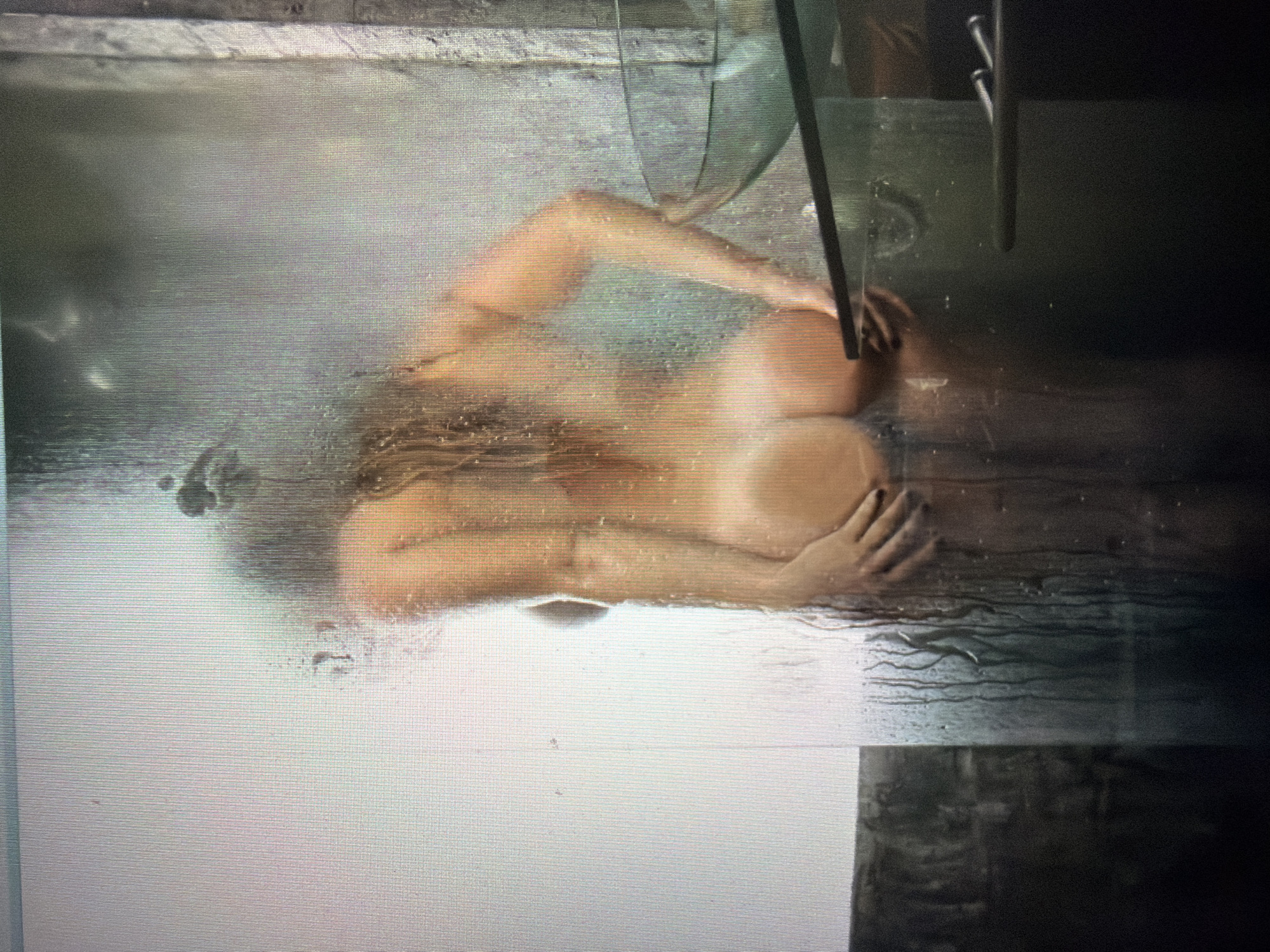 The image depicts a person lying down behind a steamy glass surface, possibly a shower door. The individual's body is mostly visible, with skin tones that suggest it is a woman. Her hair is visible, and her arms are bent, with one hand resting on her body. The background is dark and indistinct, making it difficult to discern specific objects or setting details. The steam on the glass obscures a clear view, creating a hazy effect and obscuring some of the finer details of the person's features. There is no visible text in the image. The lighting is dim, and the overall mood is intimate and private due to the obscured view and the steamy environment. The angle of the shot suggests it might be taken from a slightly elevated perspective looking down.