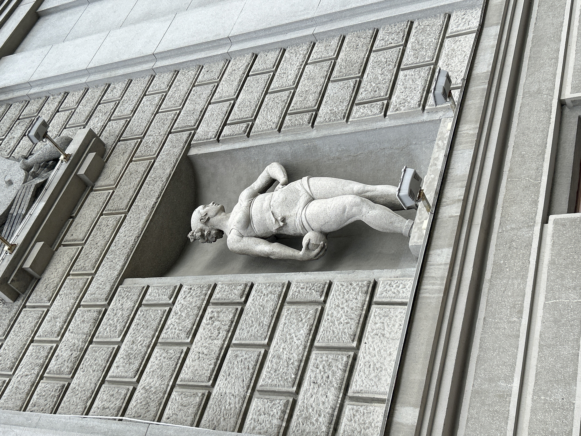The image shows a stone sculpture of a nude athletic figure holding a ball, set in a recessed niche on the facade of a building. The building's exterior is clad in stone blocks that are textured and arranged in a regular pattern. The sculpture depicts a muscular figure with short, curly hair, standing with one hand on its hip and the other holding a ball at its side. The niche is rectangular and deep, with the sculpture positioned towards the front. Two rectangular lights with grey casings are mounted on brackets below the niche, projecting outwards. The surrounding building structure is made of grey stone and features horizontal banding and recessed sections. The overall impression is of classical architecture with a modernist sculptural element. The building is likely a public or institutional building, given the scale and style of the sculpture. The location is Novokuybyshevsk, Russia.