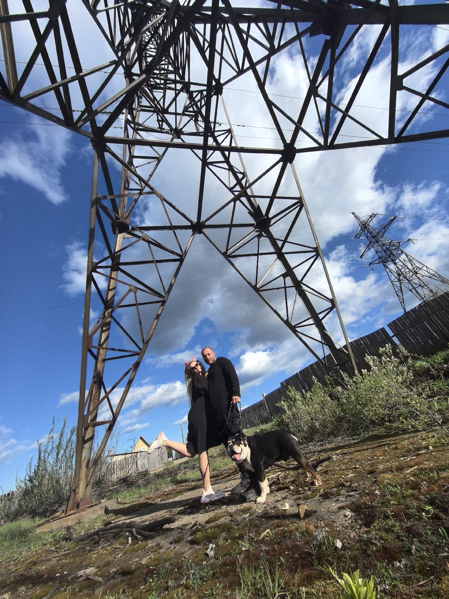 A wide-angle low-angle shot depicts a man and a woman with a dog standing below a large metal electricity pylon. Another similar structure is visible in the distance. The sky is bright blue with scattered white clouds.
The woman, positioned to the left of the pylon, has her left leg raised in a playful manner. She wears a dark dress and light-colored shoes. The man, standing next to her, is also dressed in dark clothing and holds a leash connected to the dog, which is mostly black with white markings on its chest and face.
The ground is uneven, covered in moss and sparse vegetation, with evidence of disturbed soil and roots. A wooden fence runs along the upper edge of the incline. Behind the fence, some small buildings are discernible.
The photograph was taken in the city of Noviy, Russia.