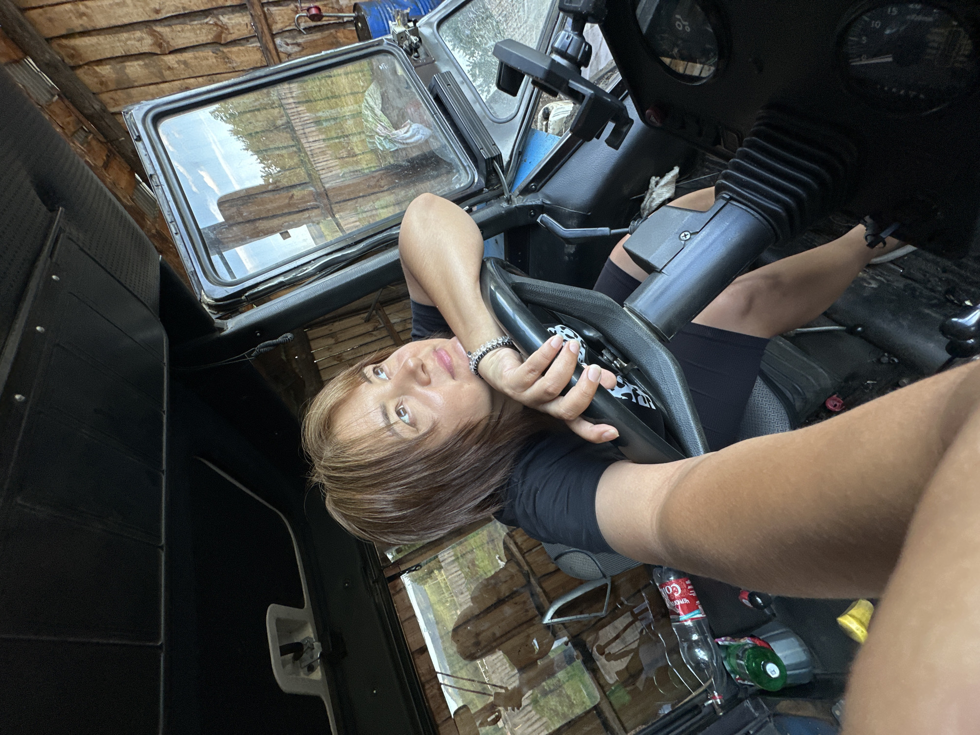A woman is sitting inside a vehicle, possibly a truck or tractor, looking out the window. She is wearing a black t-shirt and shorts. Her right arm is resting on the steering wheel. The vehicle's interior appears to be utilitarian, with a dark ceiling and worn upholstery. Through the open door and window, a rustic outdoor environment is visible, characterized by wooden structures and greenery. The overall scene suggests a rural or workshop setting.