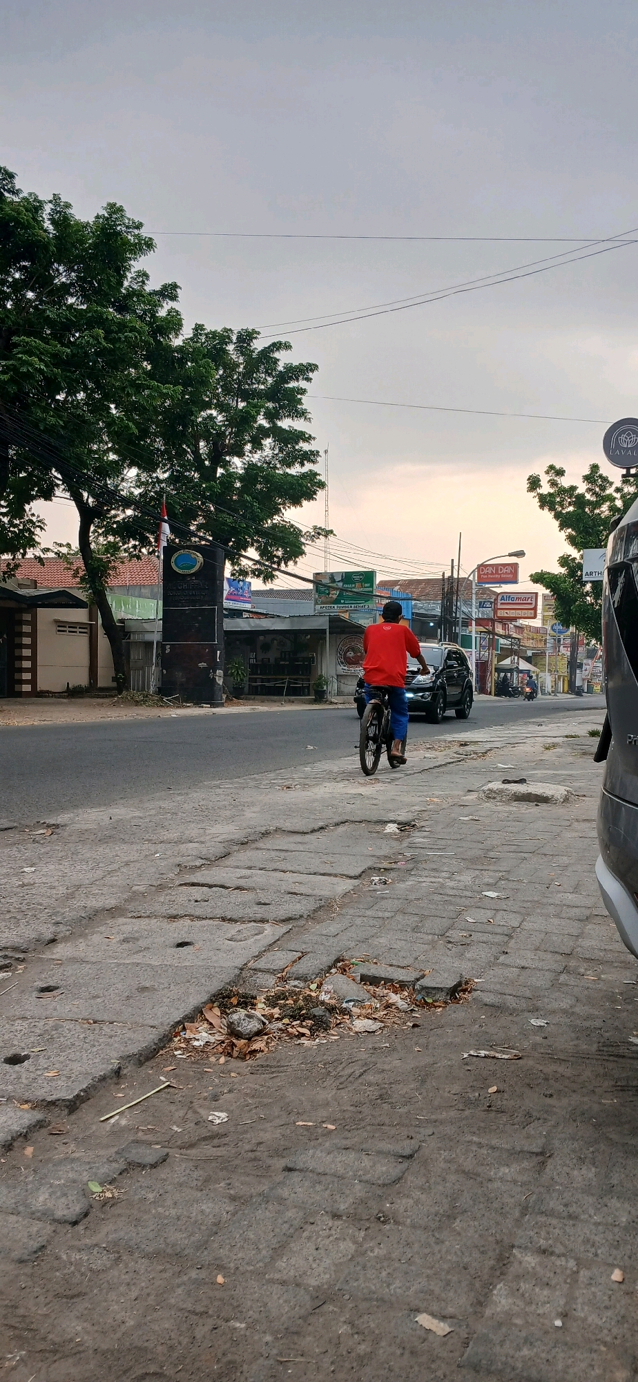 The image depicts a street scene in Cirebon, Indonesia, characterized by a paved road and a broken sidewalk. A person is riding a bicycle on the left side of the road, moving away from the viewer. The person is wearing a red shirt and blue pants. A dark-colored SUV is positioned on the right side of the road, also moving away from the viewer. The background shows several shops and buildings, including signage for "DAN DAN" and "Alfamart." Trees and utility poles are visible along the street. The sky is overcast with a hint of sunset glow. The sidewalk in the foreground is in disrepair, with cracked paving stones and debris scattered around. A large tree with dense foliage is prominent on the left side of the image, near a dark signpost with a circular logo. The overall environment suggests an urban or semi-urban setting in Indonesia.