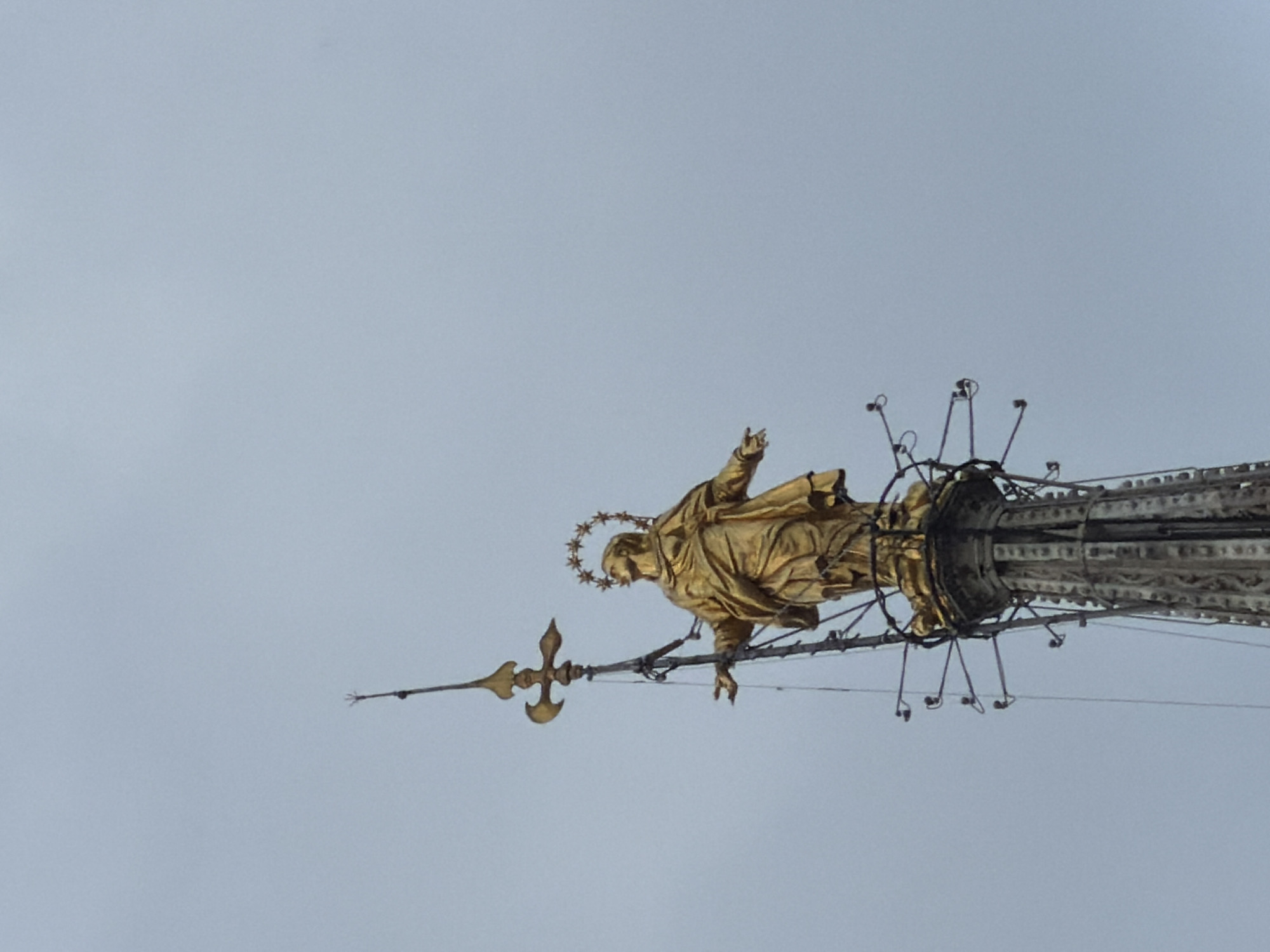 The image captures a golden statue, likely depicting the Virgin Mary given the halo of stars around her head, mounted atop a tall, ornate spire. The statue, known as the Madonnina, is a prominent feature of the Duomo di Milano (Milan Cathedral) in Milan, Italy.
The figure is rendered in a classical style, draped in flowing robes, and appears to be standing or floating with one arm raised in a gesture of blessing or welcome. Her gaze is directed upwards, and her posture conveys a sense of grace and majesty. The golden material of the statue gleams softly against the light, overcast sky.
The statue is affixed to the very peak of the cathedral's tallest spire, which features intricate architectural details and supporting structures. Below the statue, a decorative element resembling a cross or a fleur-de-lis is visible. Various wires and supports extend from the statue and the spire, possibly for structural integrity or lightning protection, and some appear to be equipped with small lights or sensors.
The sky is a uniform, pale grey, suggesting an overcast day. The image itself is rotated 90 degrees counter-clockwise, making the statue appear horizontal rather than its actual vertical orientation at the top of the cathedral. There is no visible text in the image.