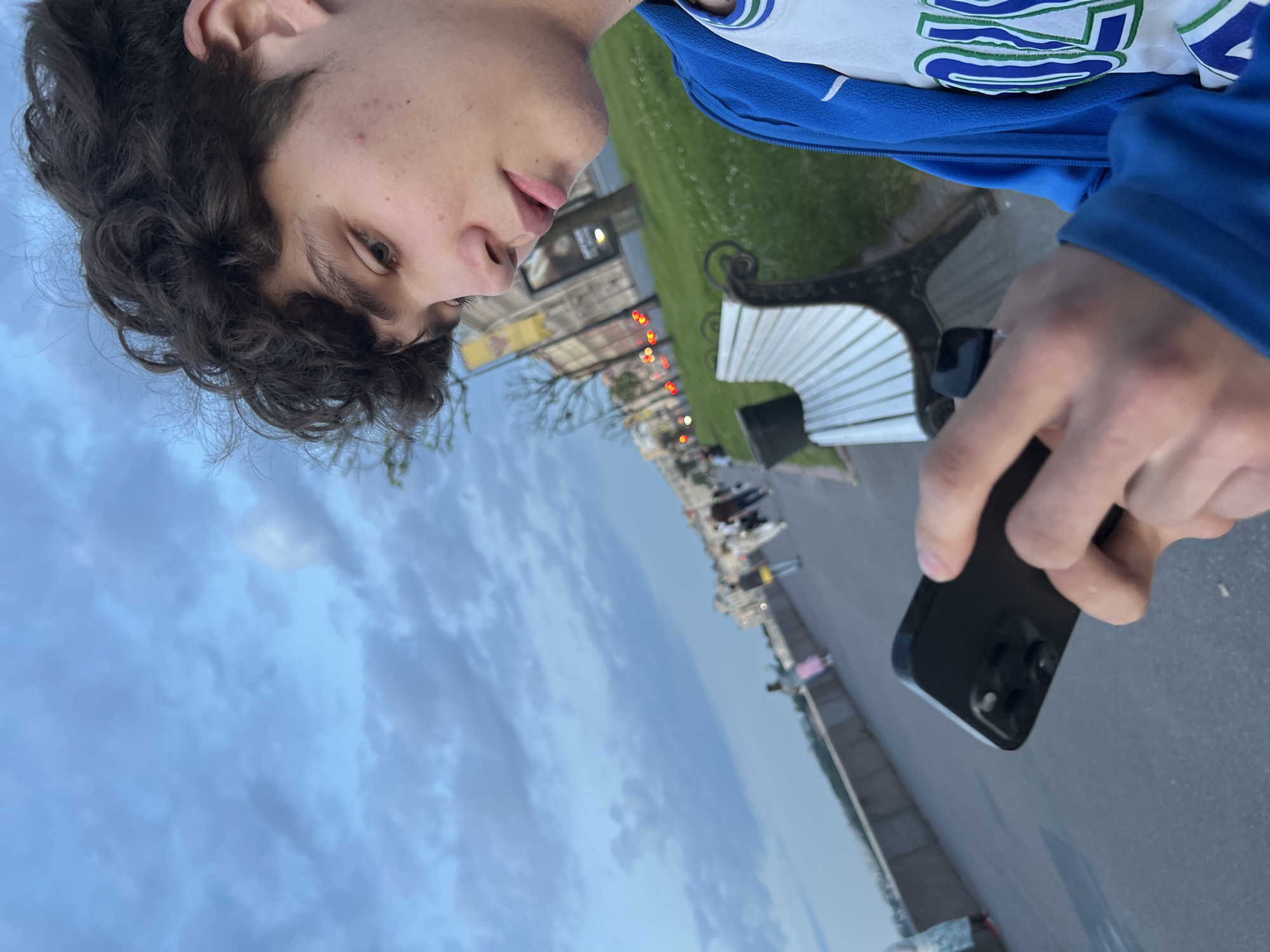 In a slightly low-angle shot taken in the city of Unknown, Russia, a young man with curly brown hair is seen looking down at his black smartphone. He is wearing a blue jacket over a white t-shirt with green and blue lettering. The man is holding the phone in his right hand, with his left hand also near it. He appears to be focused on the screen. In the background, a white park bench is visible, with a grassy area behind it. Further in the distance, several blurred figures are walking along a paved pathway next to a low wall. The sky above is cloudy and pale blue, suggesting it might be late afternoon or early evening. The overall atmosphere of the image is casual and relaxed, with the man seemingly engrossed in his device while outdoors.
