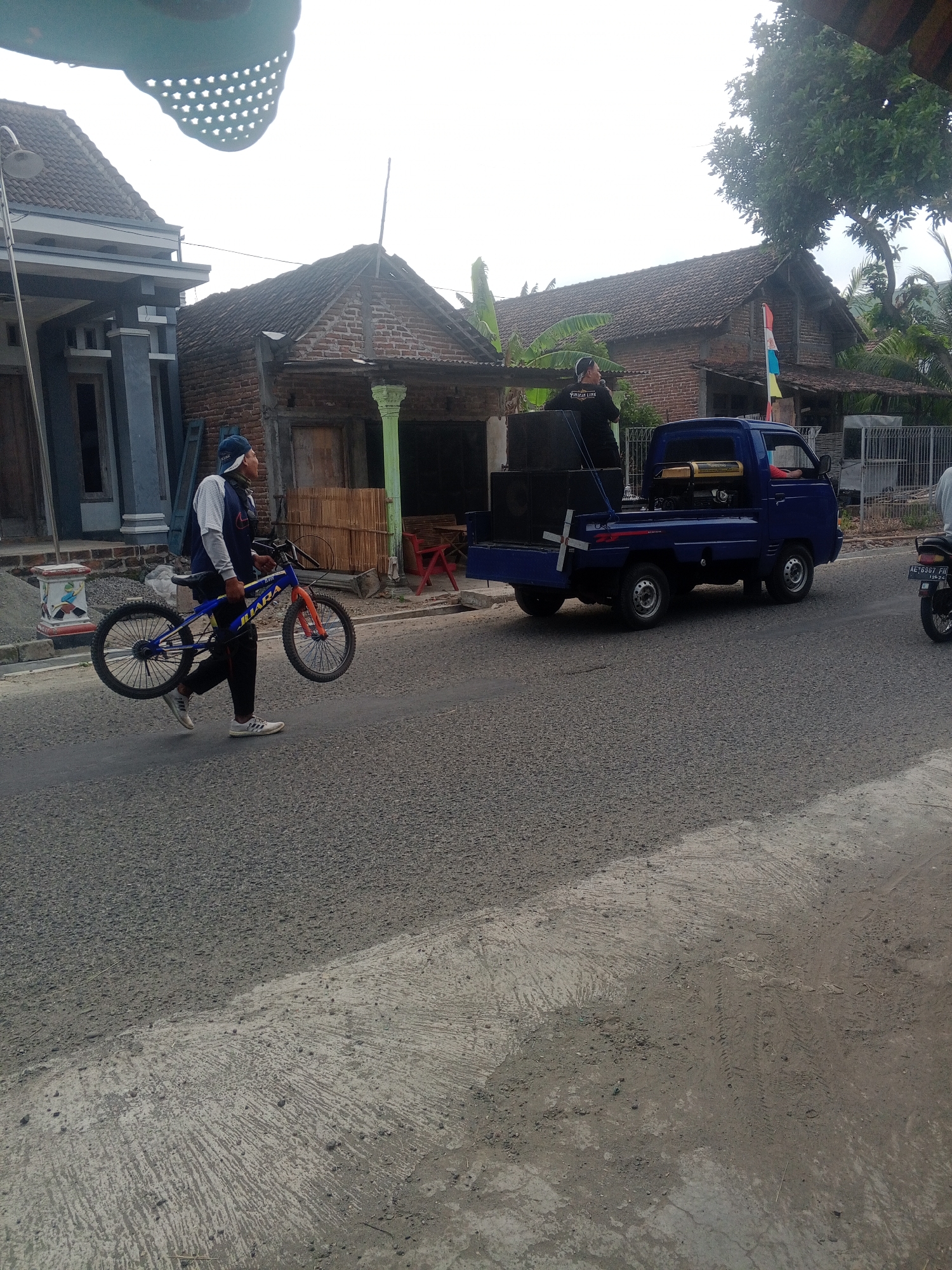 In Lembah, Indonesia, a man in a blue cap and a dark jacket carrying a blue and orange bicycle walks along a paved road. He holds the handlebars with his right hand and the seat with his left. Behind him, a blue pickup truck is parked. A person wearing a black shirt is standing in the truck bed next to large speakers. The truck has a generator and other equipment in its bed. The scene is set against a backdrop of houses with tiled roofs and lush trees. The sky is overcast.