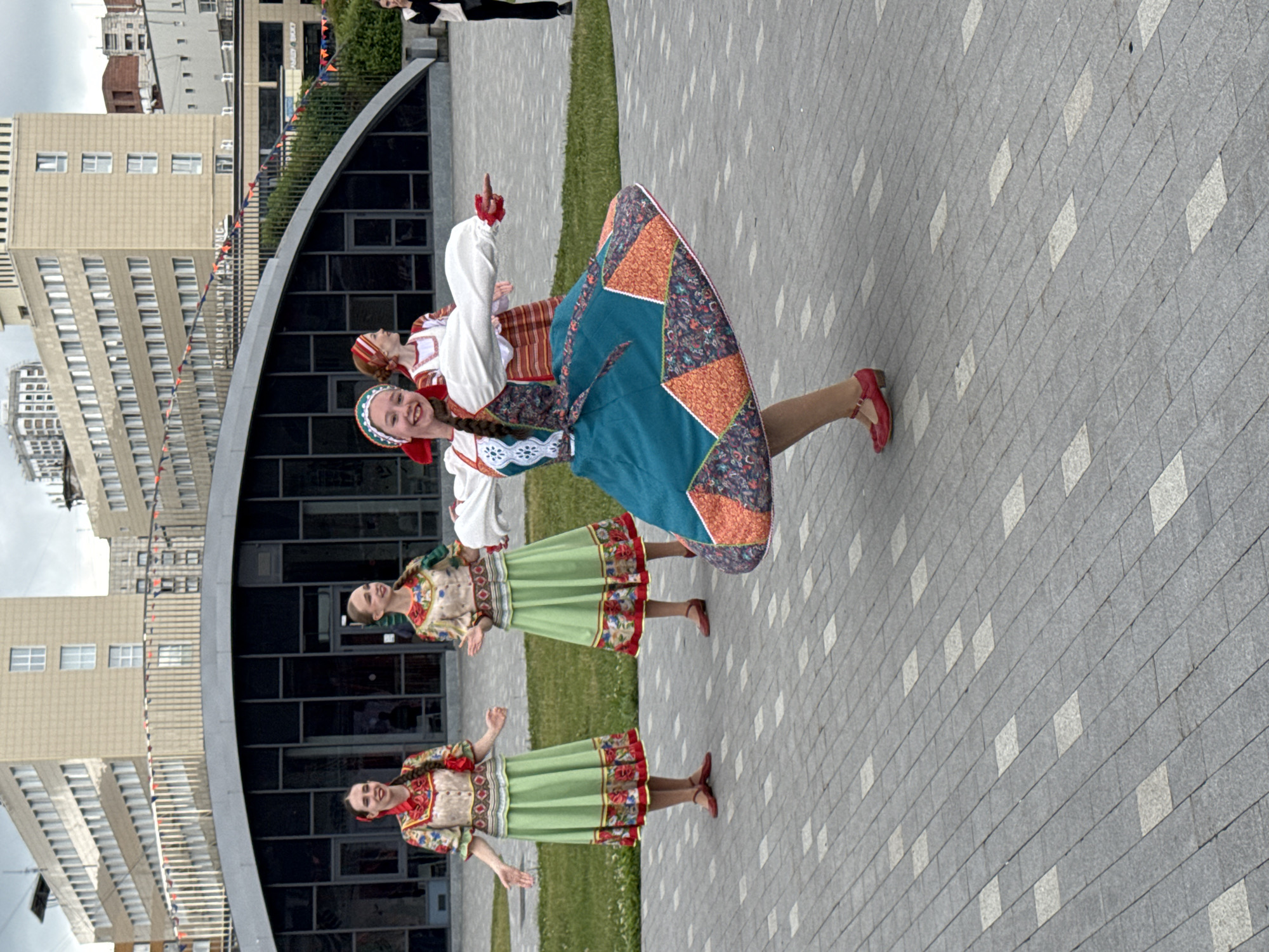 The image displays an outdoor scene in a public square or park area within the city of Perm, Russia. Four young women, dressed in vibrant traditional Russian folk costumes, are performing on a patterned paved surface under an overcast sky.
In the foreground, a young woman with a smiling expression is captured mid-dance, appearing to twirl. She wears a white long-sleeved blouse with red trim at the cuffs, a teal sarafan-style dress detailed with orange and multi-colored floral patterns, a red headband accenting her braided hair, and red Mary Jane-style shoes. Her arms are extended, and her skirt is flared due to her motion.
To her left, in the mid-ground, two other young women stand smiling and facing forward. They are identically attired in long green skirts with patterned floral borders, patterned sleeveless tops worn over white blouses, and red ribbons woven into their braided hair. Both wear red flat shoes. A fourth dancer, partially obscured, is visible behind the active dancer on the right, also in traditional attire.
The ground is composed of alternating light grey and darker grey rectangular paver blocks arranged in a grid pattern. Behind the dancers, a strip of green grass separates the paved area from background structures.
The immediate background features a modern building with a distinctive, sweeping dark-windowed facade and a curved roofline. Further behind, several multi-story buildings with light-colored facades and repetitive window structures are visible. Colorful flag bunting is strung across the background, suggesting a festive event. On the far right edge of the frame, a fifth individual, a woman in modern casual attire (black pants, white top, black vest), is observing the performance.