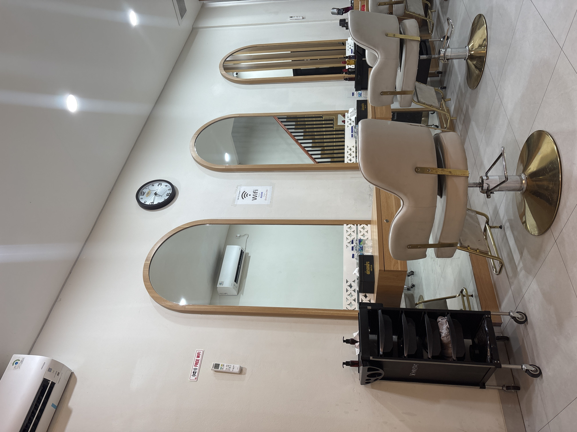 The image shows an interior view of a hair salon in North Jakarta, Indonesia. The perspective is from within the salon, focusing on three hair styling stations positioned along a wall. Each station consists of a large, arched mirror framed in light wood, a beige styling chair with a gold-colored base, and a black rolling cart for storing styling tools.
The wall behind the stations is painted a light beige color. A round clock is mounted on the wall above the middle station. Below the lowest station, an air conditioner unit is affixed to the wall. A sticker advertising free WiFi is also affixed to the wall next to a remote control.
The mirrors reflect various objects and architectural details, including a staircase and product shelving.
The floor is covered in light-colored tiles. Artificial lighting, likely fluorescent or LED, illuminates the space from the ceiling.