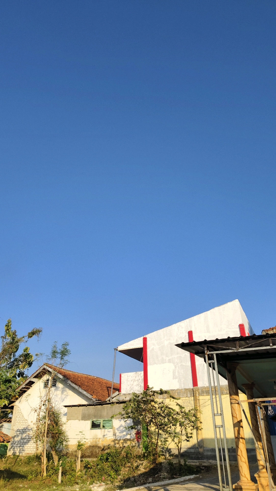 The image shows a bright, clear blue sky above a residential area in Galis, Indonesia. In the foreground, there are two houses. The house on the left has a white facade, a tiled orange roof, and a visible brick foundation. A small tree with green leaves stands in front of it. To the right of this house, a second, larger building dominates the mid-ground. This building features a white exterior with striking red vertical accents. A portion of its roof is angled, creating a geometric shape. In front of this building, there is a smaller structure with a dark roof supported by metal posts. The ground is covered with vegetation and some debris. The overall scene suggests a sunny day in a moderately developed area.