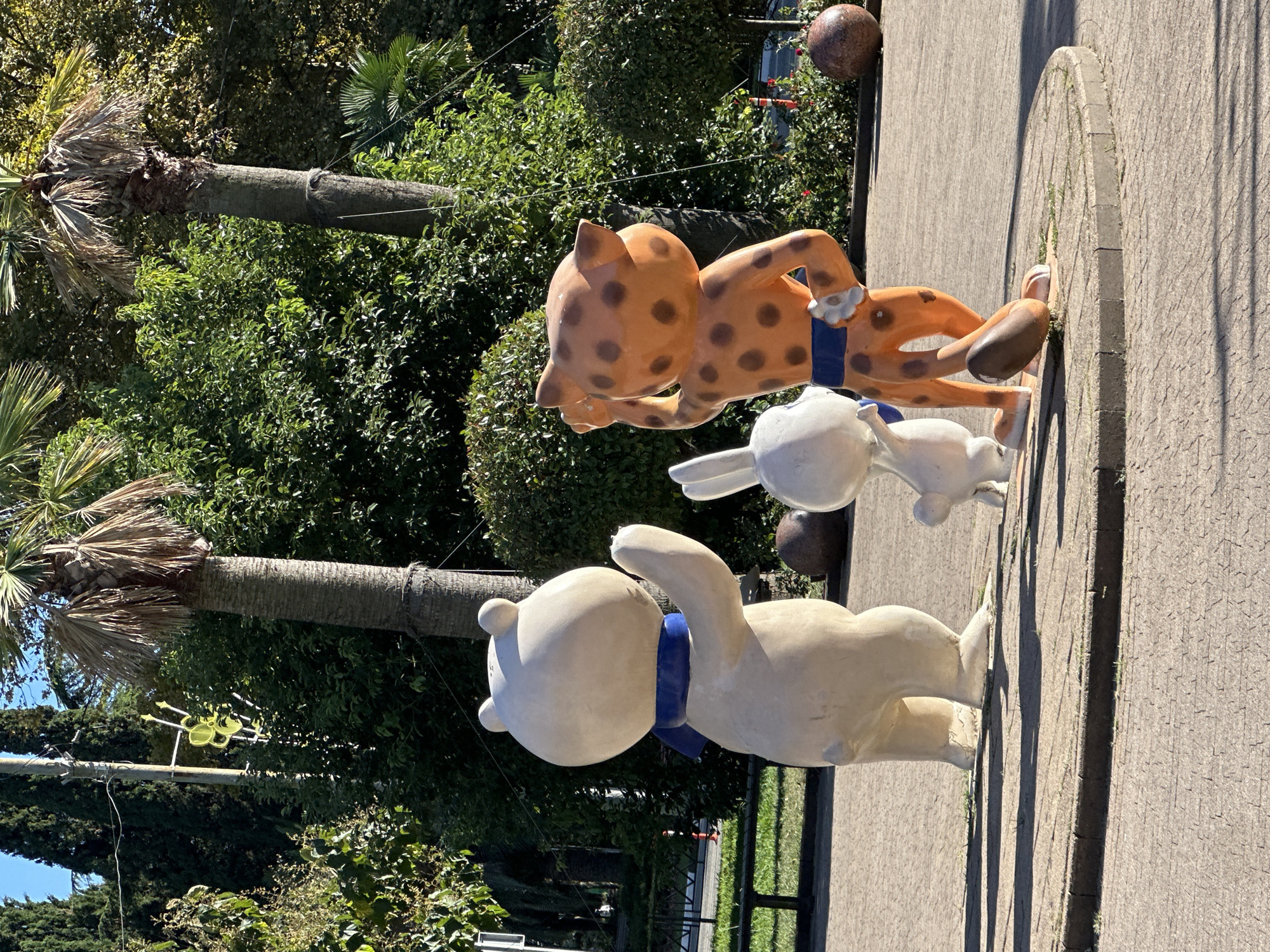 Three cartoon animal sculptures are arranged in a park setting with palm trees and lush green foliage in the background. The sculpture on the left depicts a white bear wearing a blue scarf. To its right is a smaller white rabbit sculpture. The sculpture on the far right is an orange leopard with brown spots, wearing a blue belt and white wristbands. The leopard appears to be striking a pose. All three sculptures are standing on a circular paved area. In the background, there are more palm trees and dense greenery, suggesting a warm climate. The ground is paved with a textured material, and shadows are cast by the sculptures and trees. The scene is brightly lit by sunlight. The park setting and the presence of palm trees suggest a location within Sochi, Russia, a city known for its subtropical climate and status as a resort destination.