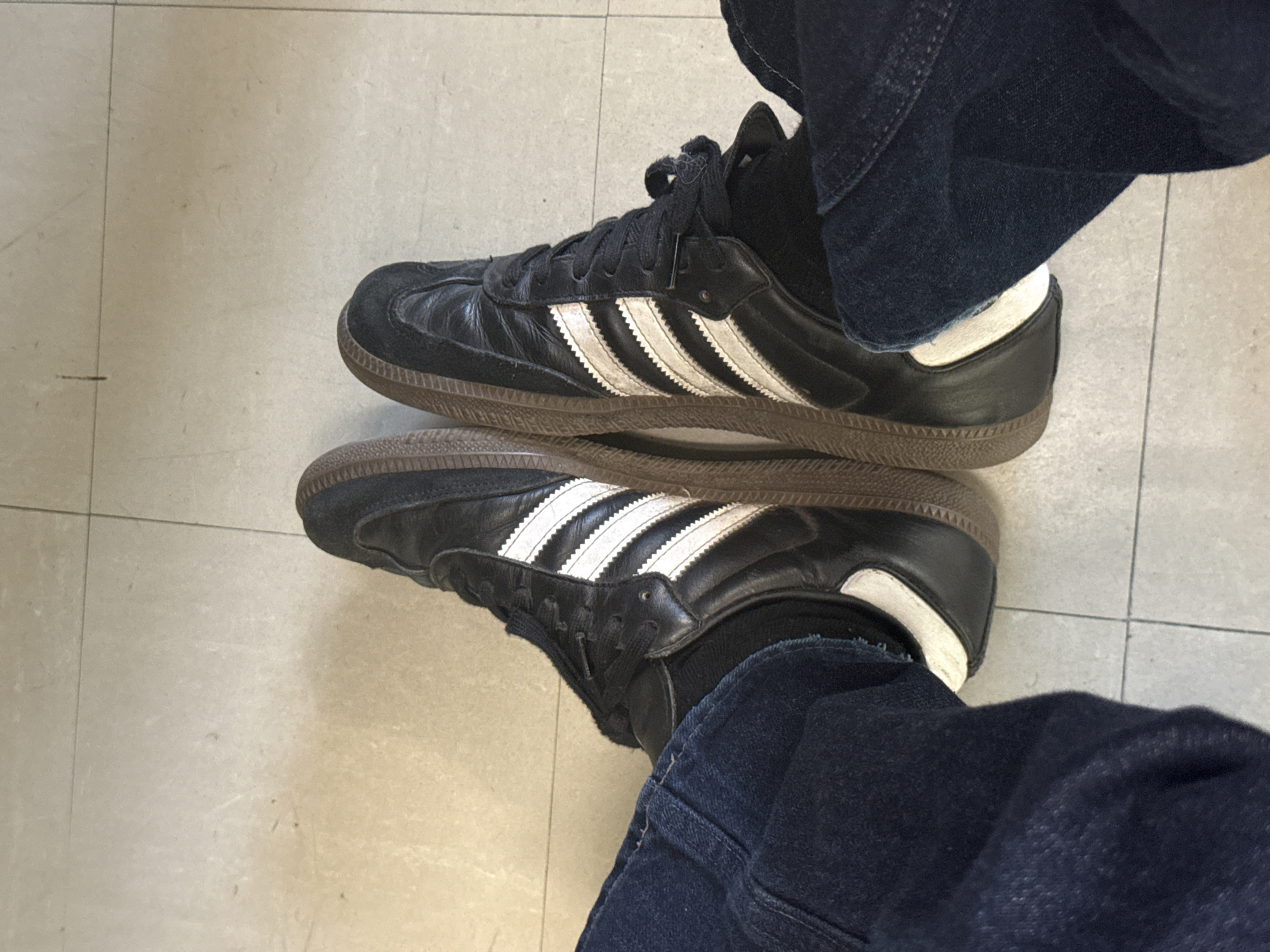 A first-person, overhead shot depicts a pair of feet wearing black Adidas Samba sneakers, dark-colored socks, and dark-wash denim jeans. The person is standing on a light-colored, tiled floor. The sneakers are positioned closely together, parallel to each other. There is no visible interaction or action taking place in the image. The location is not discernible from the image.