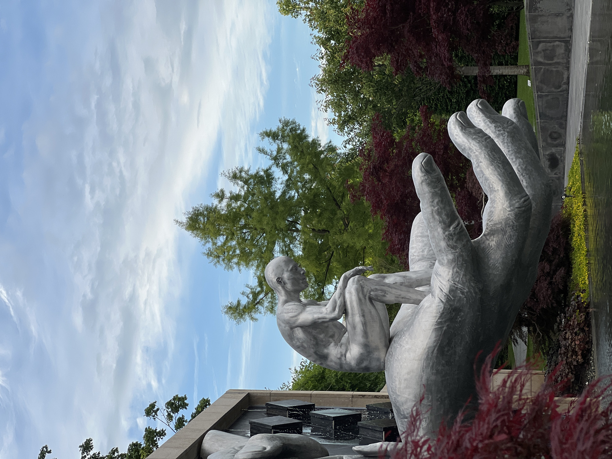 This image captures a striking outdoor sculpture in a landscaped park setting. The central focus is a large, metallic grey sculpture featuring a bald, lean human figure seated in a contemplative pose, with knees drawn to its chest. This figure rests within the palm of an enormous, intricately detailed hand, also rendered in a metallic grey finish, showing distinct fingers, knuckles, and fingernails.
Below the sculpture, a modern water feature can be seen, consisting of dark, rectangular blocks arranged in steps from which water appears to be flowing or cascading. The water surface is dark and reflective. Vibrant red-leafed foliage partially frames the lower right foreground, while more dark red-leafed trees are visible in the mid-ground on the right.
The background is filled with lush greenery, including tall, verdant trees, suggesting a natural and well-maintained environment. The sky above is a clear blue with scattered white, wispy clouds, indicating a bright daytime setting, likely morning or afternoon. The overall atmosphere is serene and artistic. No discernible text is visible in the image.