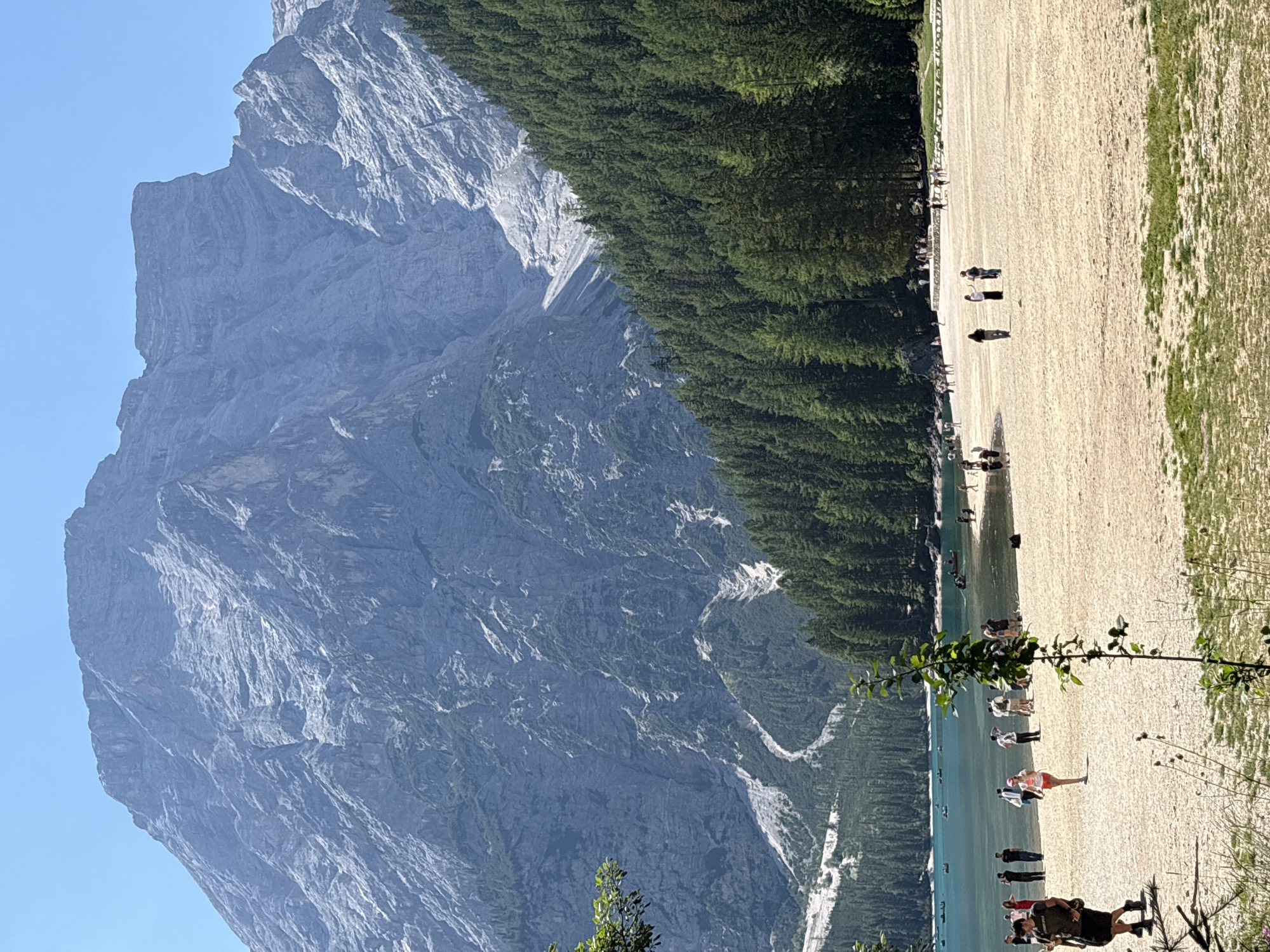 The image captures a scenic view of a mountainous landscape with a lake and a sandy beach. A large, rugged mountain dominates the background, with steep, rocky slopes and a jagged peak. The mountain is partially covered in vegetation, with a dense forest of coniferous trees ascending its lower slopes. A body of turquoise water, likely a lake, is visible in the mid-ground, with several small boats on its surface. The foreground features a wide, sandy beach where numerous people are scattered. Some individuals are walking, while others are standing or gathered in small groups. The overall scene suggests a popular tourist destination, likely in the Italian Dolomites.