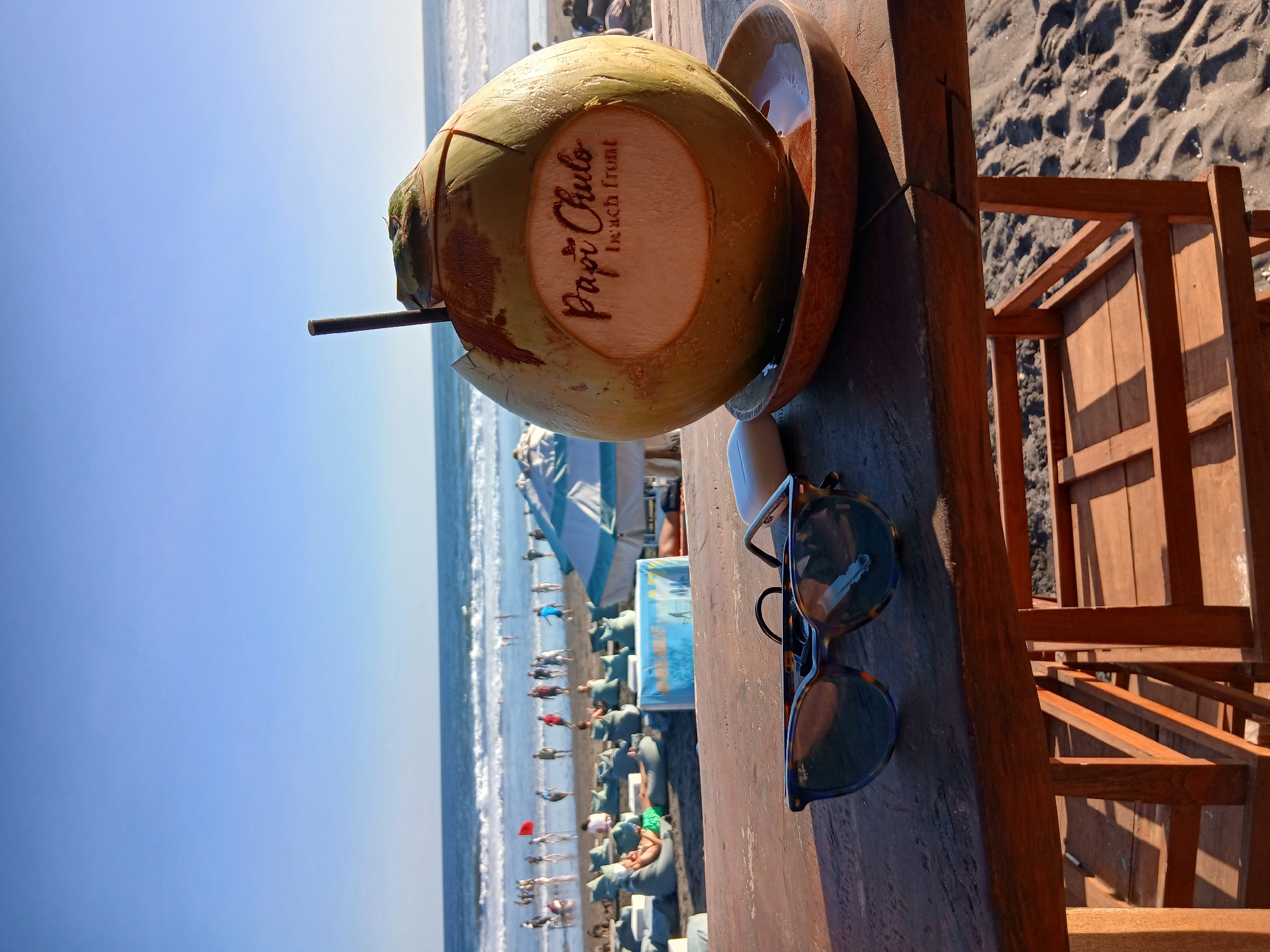 An eye-level outdoor shot captures a refreshing coconut drink with a straw and the logo "Papi Chulo beach front" carved into it, sitting on a wooden saucer atop a wooden table. To the left of the coconut, a pair of tortoise-shell sunglasses rests next to a white earbud case. The table is positioned on a sandy beach, with the ocean waves and a clear blue sky forming the background. In the middle ground, several people are visible on the beach, some near the water's edge and others lounging on beanbag chairs under umbrellas. The overall scene suggests a relaxed atmosphere at a beachside establishment in North Kuta, Indonesia.