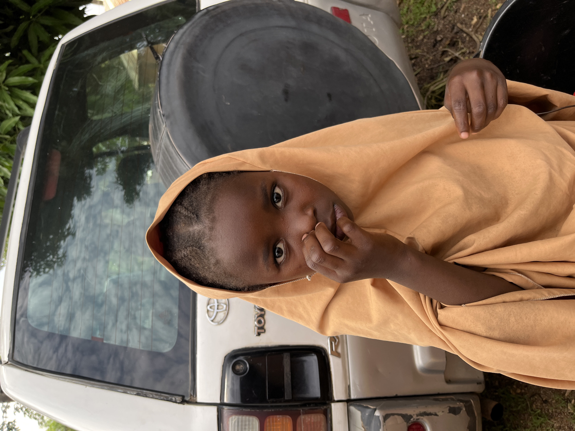 A girl with dark skin and short braids is wrapped in a tan cloth hood, looking at the camera with her hand near her mouth. She is standing near the back of a silver Toyota SUV in Ganawuri, Nigeria. The SUV has a spare tire mounted on the back. A black object is visible to the right of the girl. Green foliage is visible in the background.