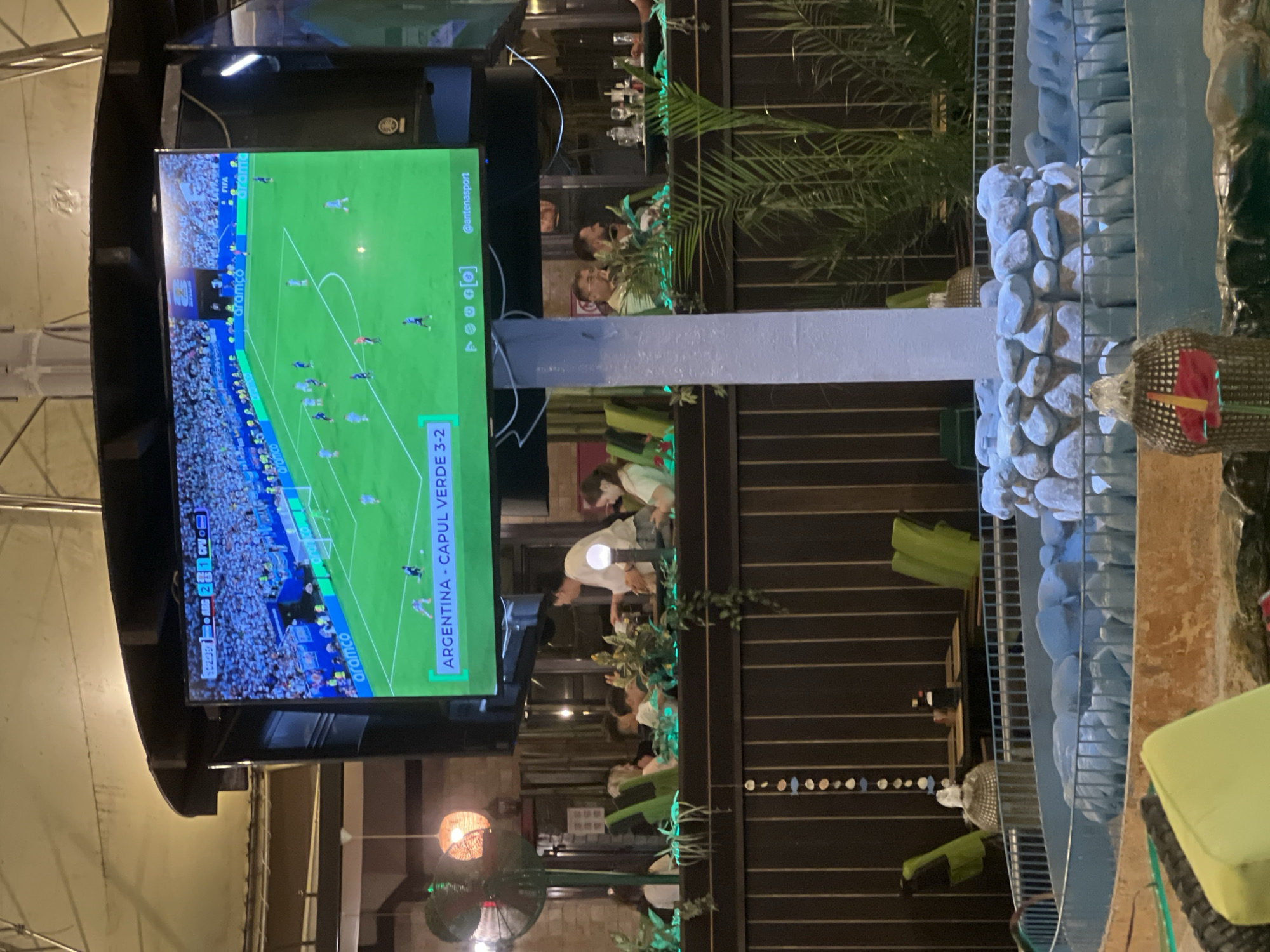 The image captures an outdoor seating area with a large television screen displaying a soccer match. The match is identified as "ARGENTINA - CAPUL VERDE 3-2," with the time "22:46" visible on the screen. The screen also shows broadcast information including "@antenasport." In the foreground, a decorative fountain with blue-tinted water and rocks is visible. Several people are seated at tables in the background, some appearing to be dining or socializing. Green chairs and tables are present in the seating area, interspersed with potted plants. A ceiling fan is also visible on the left side of the image. The overall atmosphere suggests a casual dining or entertainment venue. The provided context indicates the location is Constanța, Romania.