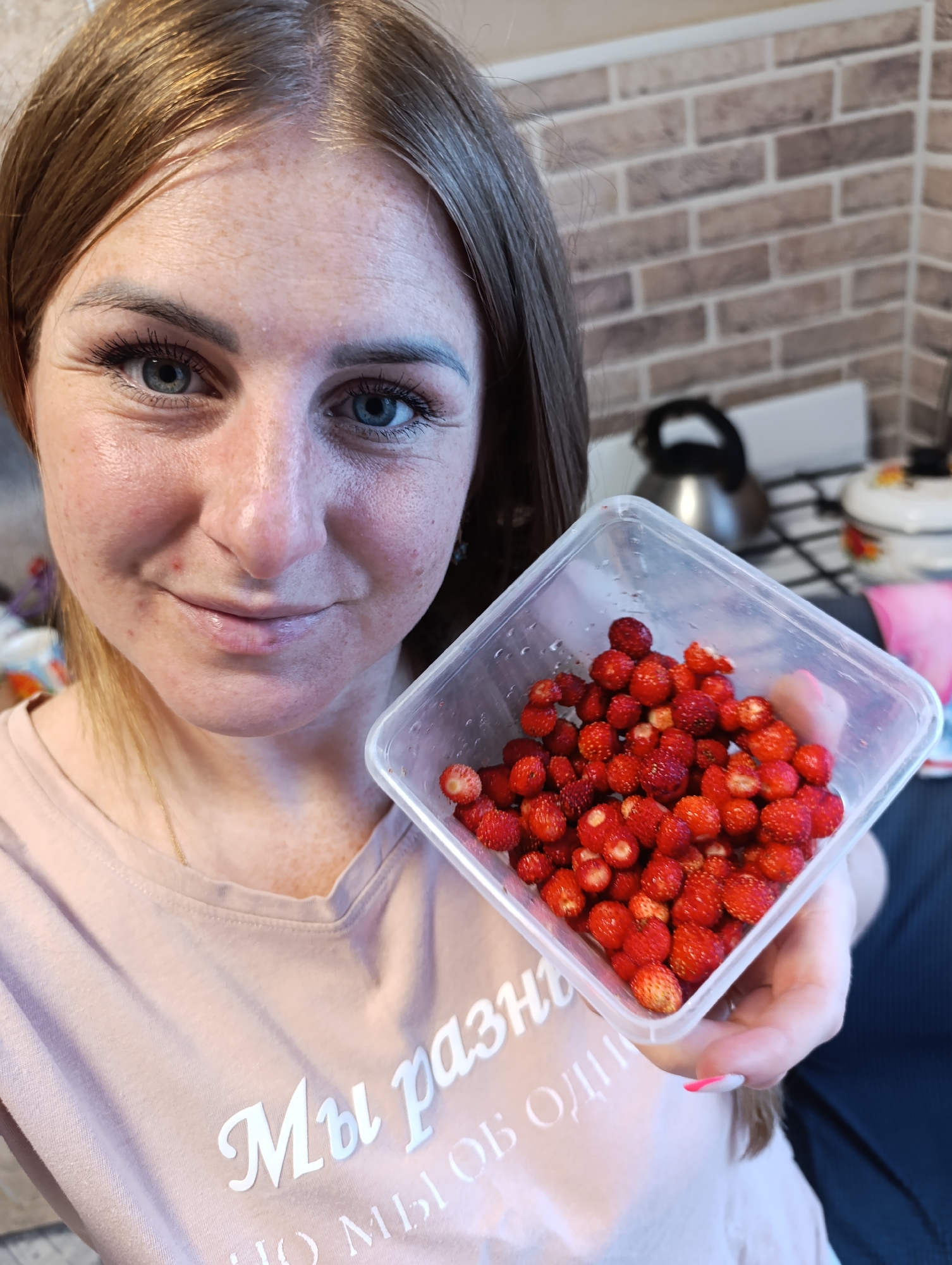 A young woman with light brown hair and blue eyes is holding a clear plastic container filled with wild strawberries. She is wearing a light pink t-shirt with white text. Her skin is fair and freckled. She is looking towards the camera with a slight smile. The background appears to be a kitchen with brick tile walls and a stove. The overall setting suggests a domestic environment.