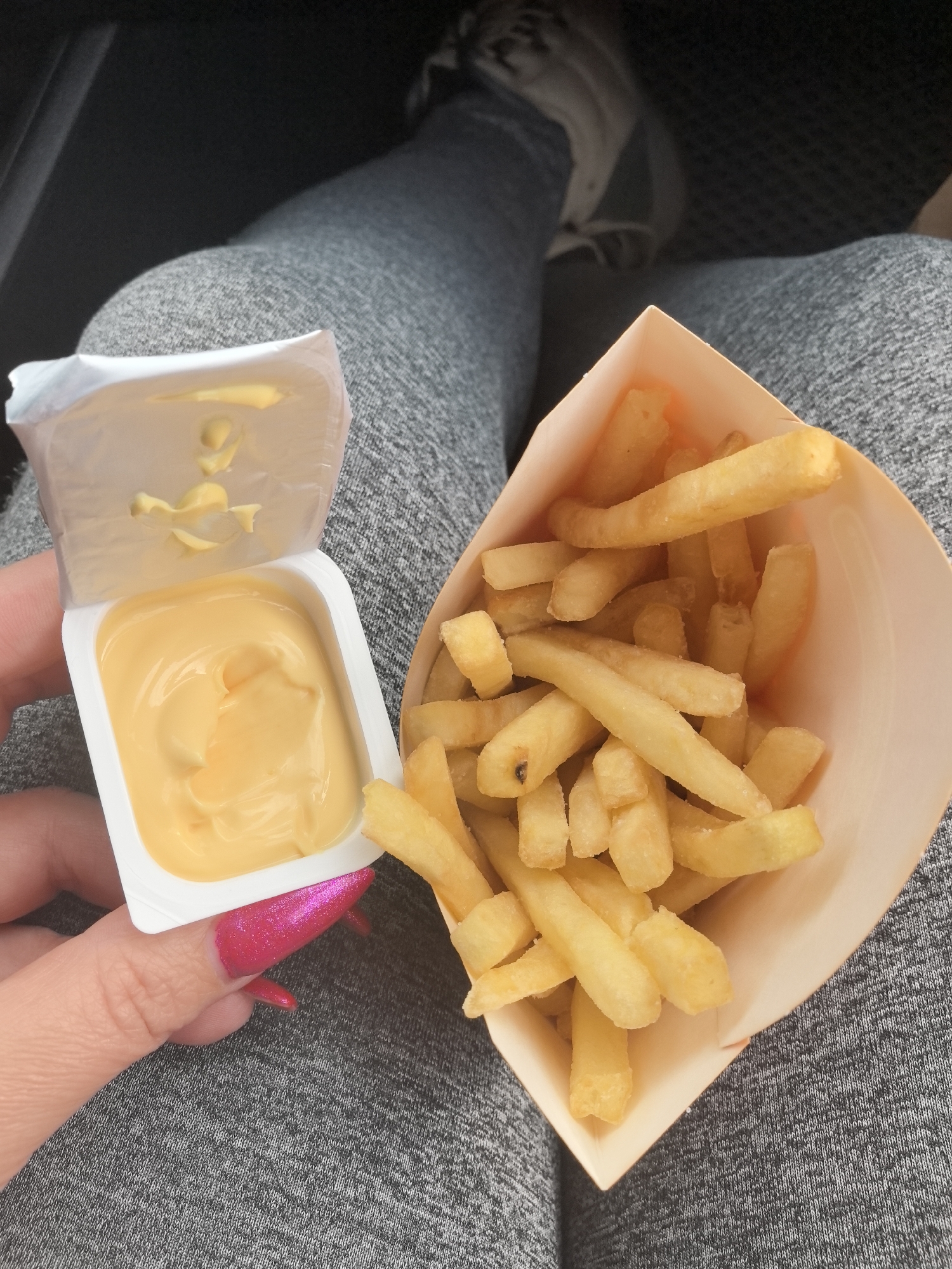 A person is holding a container of cheese sauce and a container of french fries in their lap. The person is wearing gray sweatpants and gray sneakers. The photograph was taken in an indoor setting, possibly a car. It is not possible to determine if the location is in the city of Kstovo, Russia from the image alone.