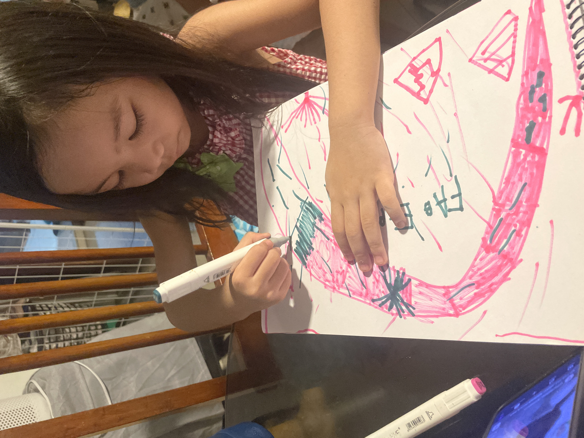 In this image, a young girl with dark hair is seated at a table, engrossed in drawing on a white sheet of paper with pink and green markers. Her right hand, holding a marker, is positioned over the paper, actively creating the drawing. Another marker lies on the table to her left, near a tablet device displaying a bright blue screen. The background shows wooden chairs and a glimpse of what appears to be an indoor setting. The girl is wearing a red and white checkered top. This scene is likely located in a residential area of Davao City, Philippines, given the general context of such images.