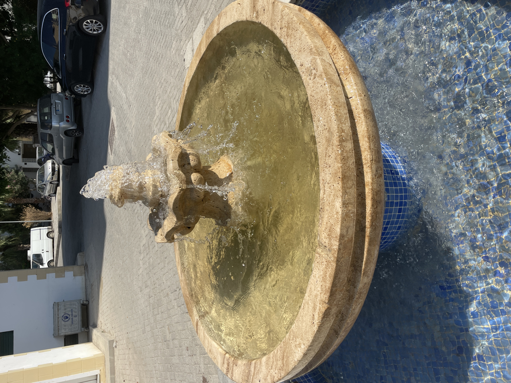 A stone fountain with water actively flowing is the central subject of this outdoor scene, situated on a paved surface in Trikomo, Northern Cyprus. The fountain has a tiered design, with a lower basin made of beige stone and a smaller, multi-tiered structure at the top from which water cascades. The water in the basin is a murky yellow-brown color, with ripples and splashes clearly visible. The base of the fountain is surrounded by a tiled pool, featuring small blue square tiles.
In the background, a white building with yellow trim is partially visible on the left. Adjacent to the building is a large, white rectangular dumpster with wheels, bearing a blue logo and text that appears to read "CAESAR PROJECTS". Further back, a white van is parked. The paved ground is made of interlocking grey bricks, and palm trees are visible in the middle ground and background, suggesting a warm climate. Several cars are parked on the street behind the fountain, including a silver hatchback, a silver minivan, and a black sedan. The sky is clear and bright, indicating a sunny day. The scene appears to be a public area, likely in a town or city.