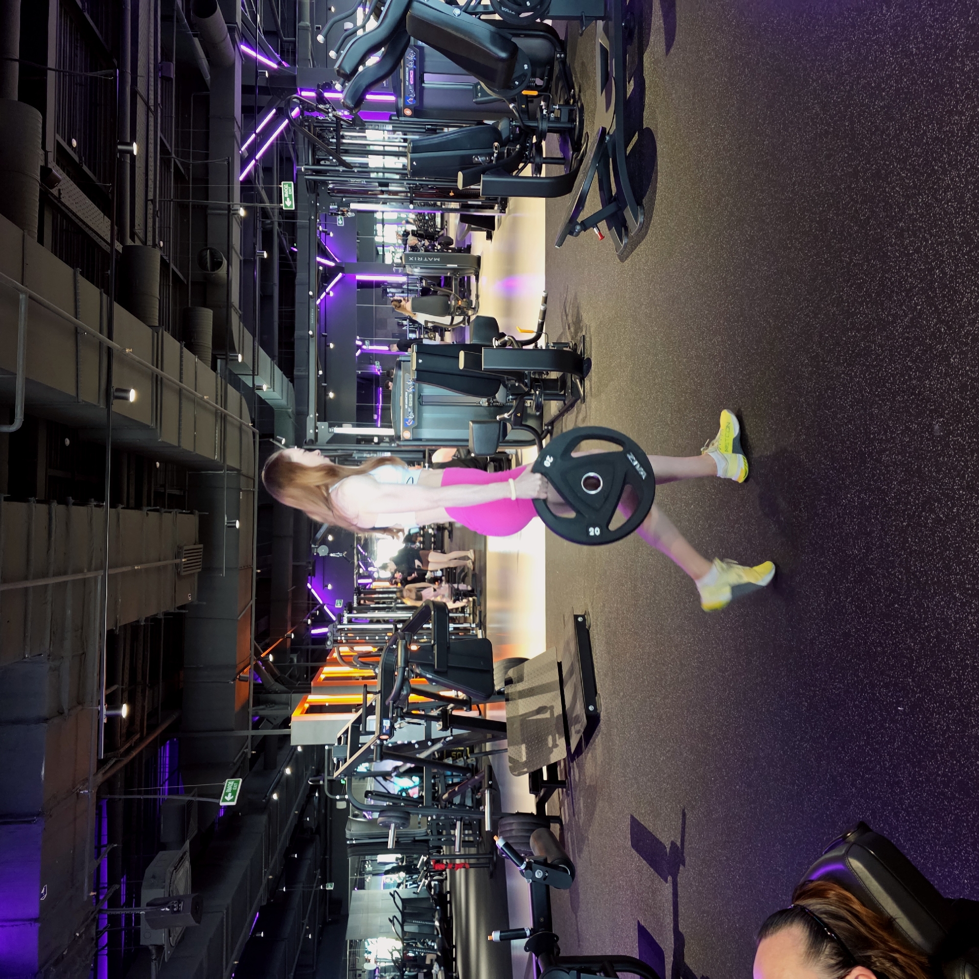 The image depicts a modern, well-equipped gym with an industrial design aesthetic. The main subject is a woman in the mid-ground, positioned facing right, engaged in a workout. She has long, light brown hair and is wearing a light-colored sports bra or crop top paired with bright pink shorts and striking yellow sneakers. She is actively holding and lifting a black "ZIVA" branded 20kg barbell weight plate.
The gym features exposed dark grey ductwork, pipes, and structural elements on the ceiling and walls, contrasted by vibrant purple and orange-yellow LED strip lighting that creates a dynamic and modern atmosphere. The floor is a dark, speckled rubberized material.
In the bottom right corner, partially visible, is another woman with dark hair tied back, wearing a headband, appearing to be seated on a piece of gym equipment. Reflections of other gym-goers and exercise machines, some bearing the "MATRIX" brand, can be seen in the background, suggesting a busy environment. A green "EXIT" sign with an arrow pointing right is visible, featuring both English "EXIT" and Cyrillic "ВЫХОД" text, consistent with the Saint Petersburg, Russia location context. The overall scene suggests a focused and energetic fitness activity in a stylish, contemporary gym setting.