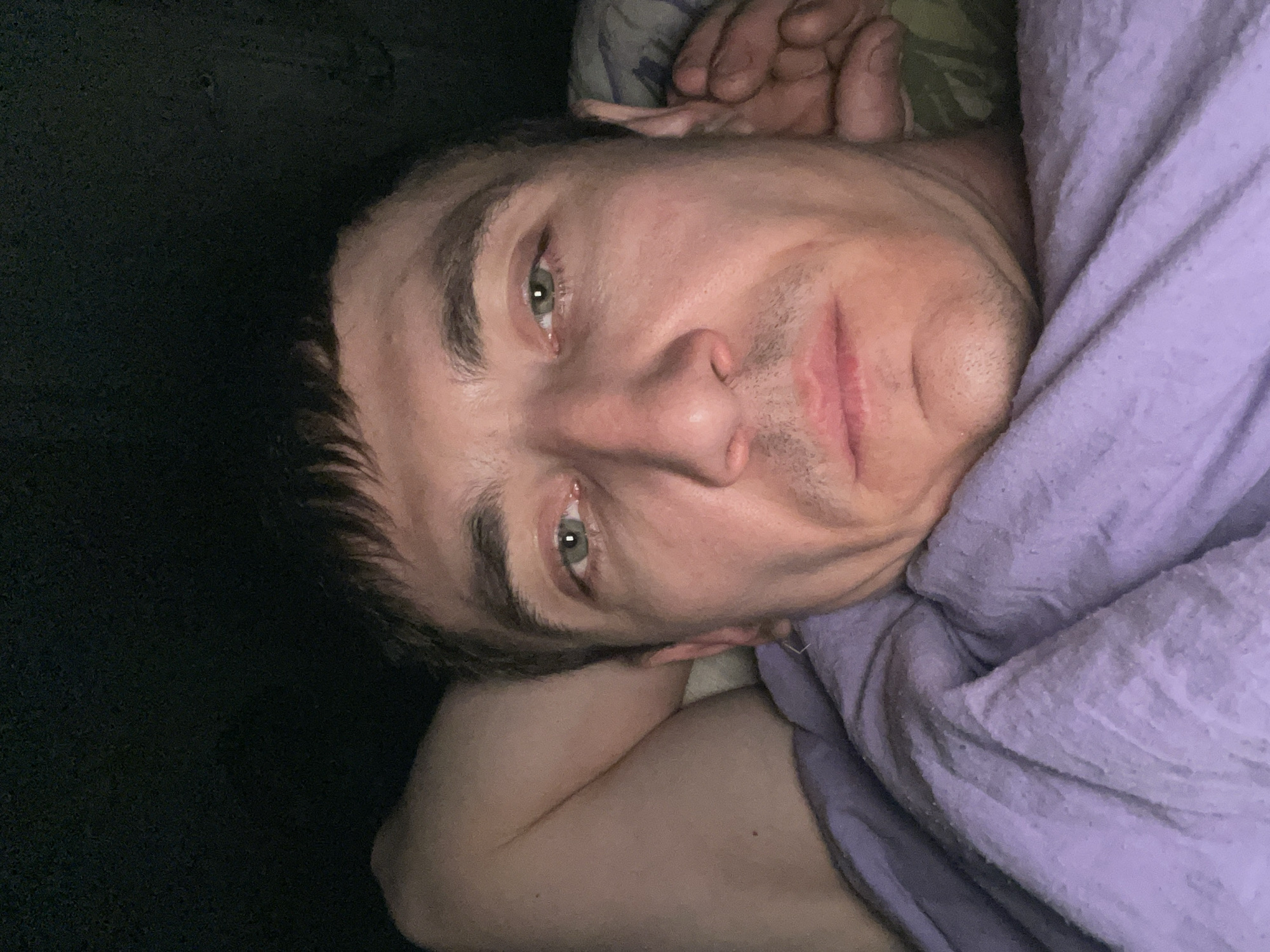 A close-up, low-angle, waist-up selfie depicts a Caucasian male lying in bed, facing the camera. He has short, dark brown hair and light green eyes. He is wearing a purple blanket. The background is dark and appears to be indoors, possibly in a bedroom in Laishevo, Russia. The lighting is dim, creating a somber mood. The man's left arm is bent behind his head, and his right hand is near his ear. His expression is neutral.