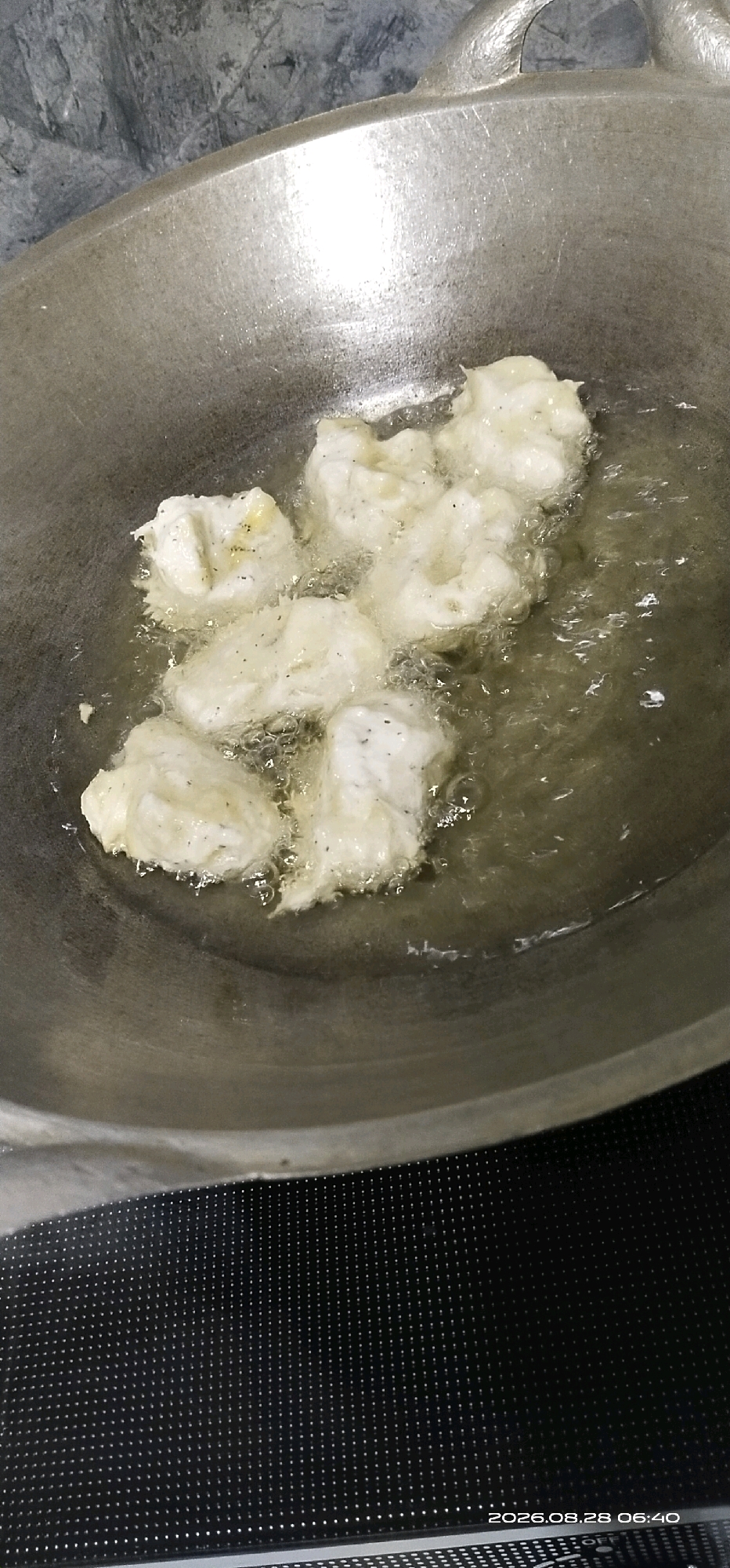 The image shows a close-up, overhead view of several pieces of light-colored, irregular-shaped food items, likely dough-based, being deep-fried in a wok. The food is submerged in hot, clear oil, and bubbles are visible around the pieces, indicating the frying process. The wok is made of a dull metal, possibly aluminum or stainless steel, and has a rounded bottom and two handles on the sides, one of which is partially visible. The background is out of focus, showing a textured, possibly stone-like surface above and a dark, dotted surface below, which appears to be the stovetop. A timestamp "2026.08.28 06:40" is visible in the bottom right corner of the image. The scene is likely a kitchen in Matabu, Indonesia, where such food preparation is common.