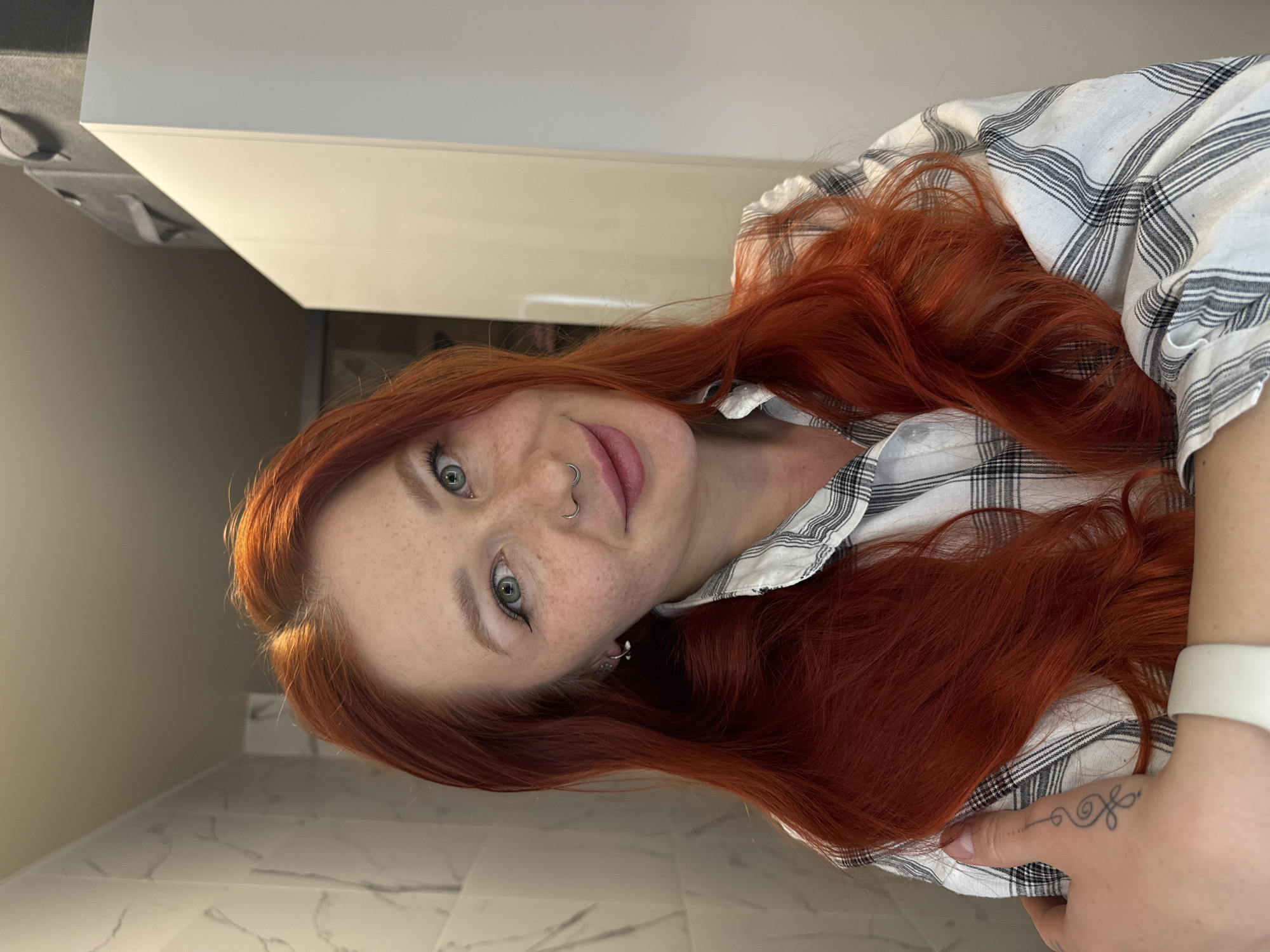 A woman with red hair, freckles, and green eyes looks directly at the camera. She has a nose ring and is wearing a white and gray plaid shirt. Her left hand is visible, adorned with a tattoo. She is located indoors, with a white marble-patterned surface beneath her. In the background is a white cabinet and a wall. The location is not Rostov-on-Don, Russia.