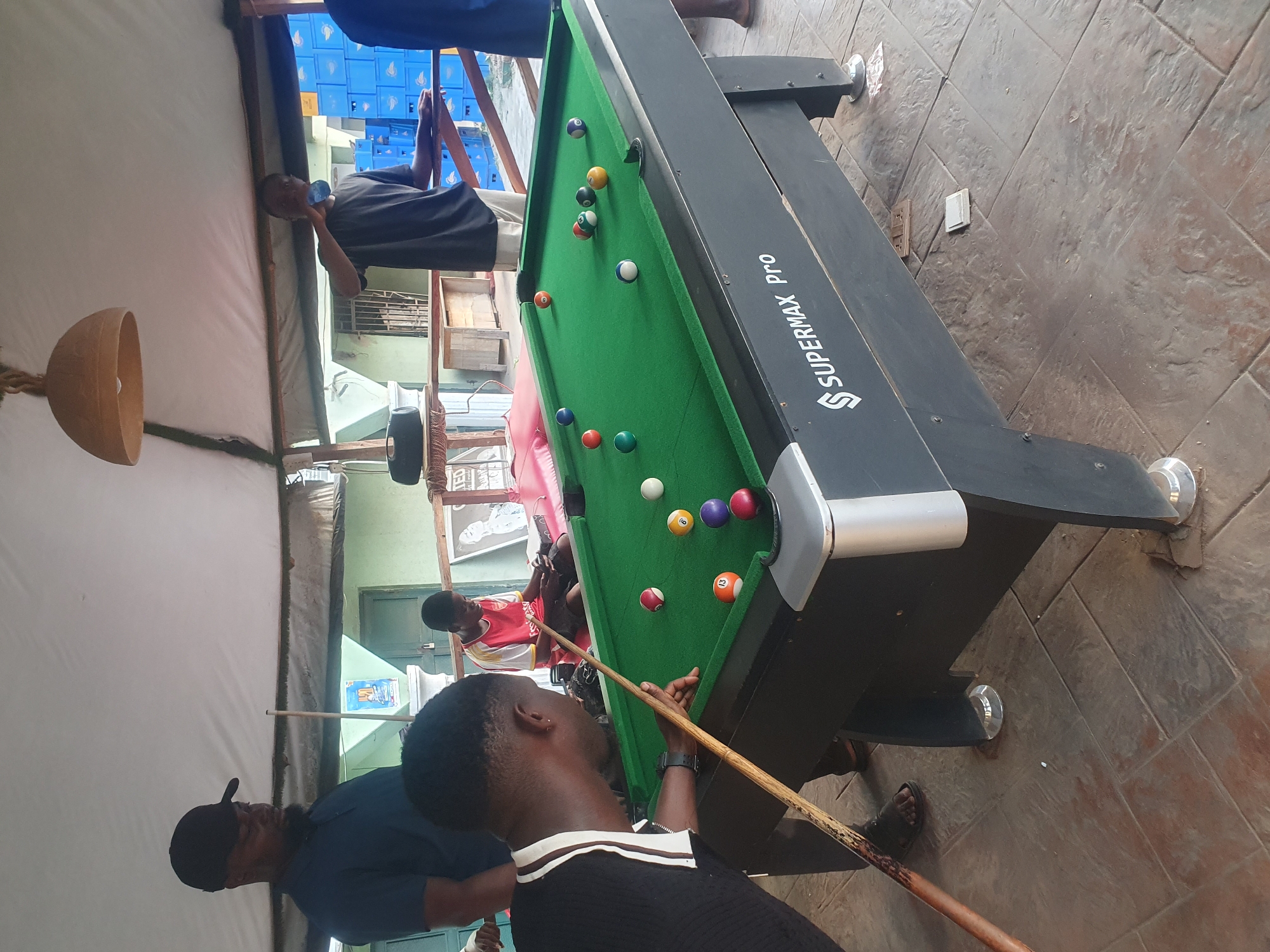 In Lokoja, Nigeria, a group of men are playing pool on a "SUPERMAX Pro" brand pool table. The pool table has a green playing surface and multiple colored pool balls are visible. One man is holding a pool cue and preparing to take a shot. Other men are observing the game. The setting appears to be an outdoor or semi-outdoor area with a covered ceiling and textured floor. Blue crates can be seen in the background. A woven lamp hangs from the ceiling.