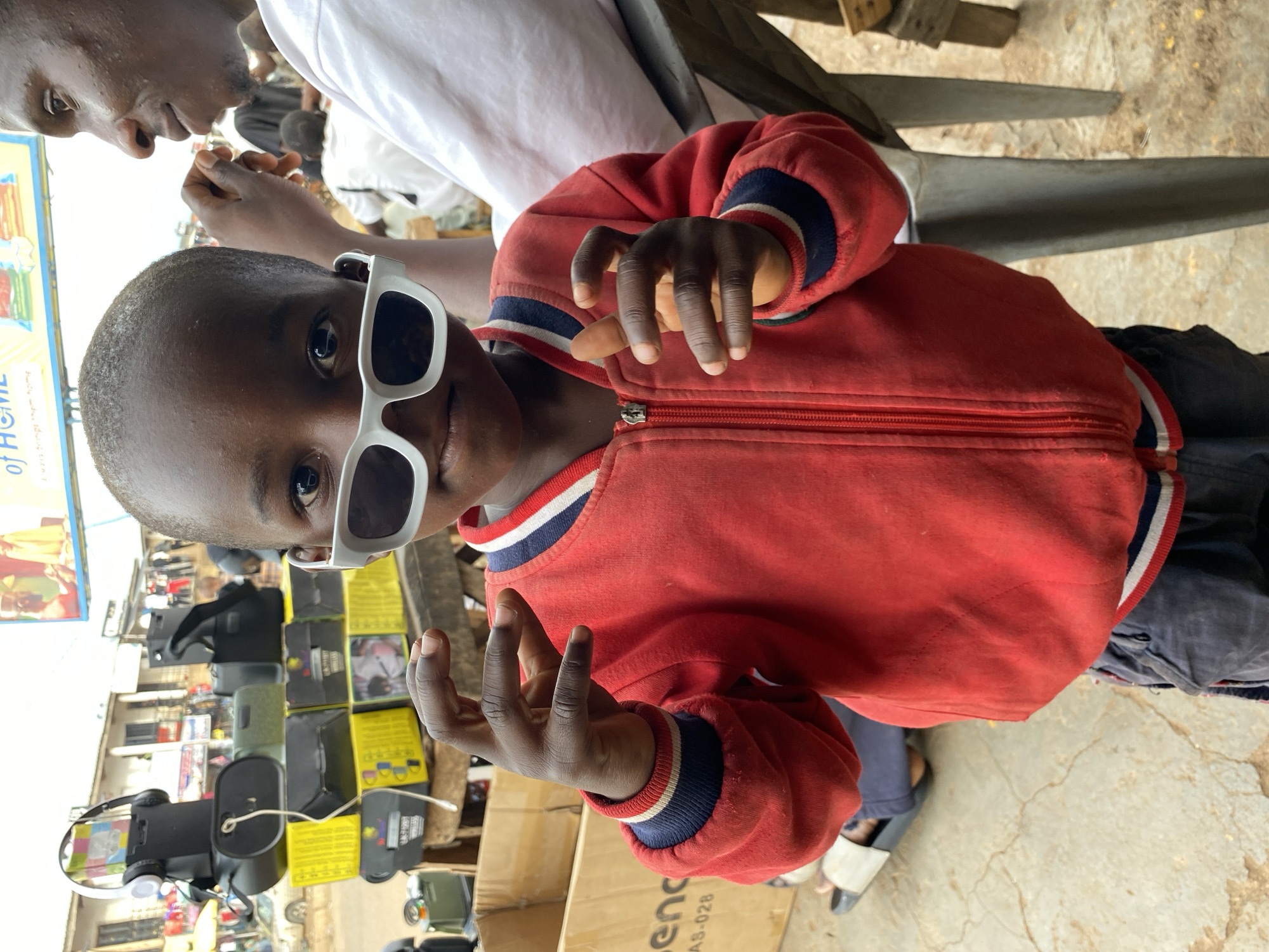 The image shows a child wearing white sunglasses and a red jacket with blue and white striped cuffs and collar. The child is positioned in the center of the frame, looking towards the camera and gesturing with both hands. An adult partially visible in the top of the frame is wearing a white shirt. The background shows a vendor's display with various items for sale, including boxes, headphones, and electronic equipment. The location is Bukuru, Nigeria, as evidenced by the local market scene and surrounding environment. The ground is a light-colored, cracked surface.