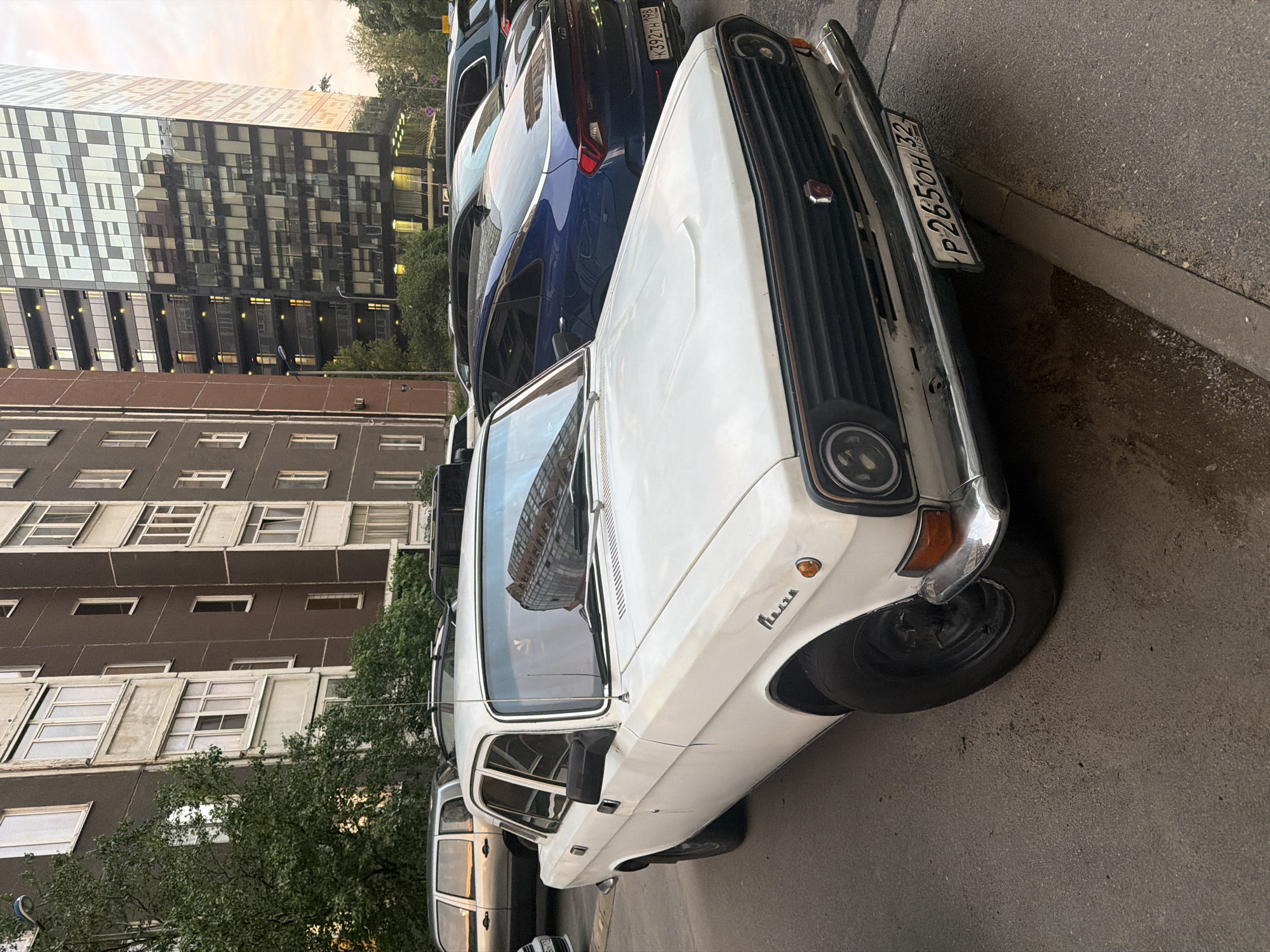 A white, vintage sedan is parked on an asphalt surface in front of apartment buildings and a modern glass-facade high-rise building. The car, which appears to be a Volga, is positioned at an angle, with its front end closest to the viewer. It has a chrome grille, round headlights, and a license plate with the characters "P 265OH 32" and "RUS". Reflections of the modern building are visible on the car's windshield and hood. Other vehicles, including a dark blue sedan and a silver station wagon, are parked nearby. Trees and greenery are visible between the buildings. The sky suggests it is either late afternoon or early evening.