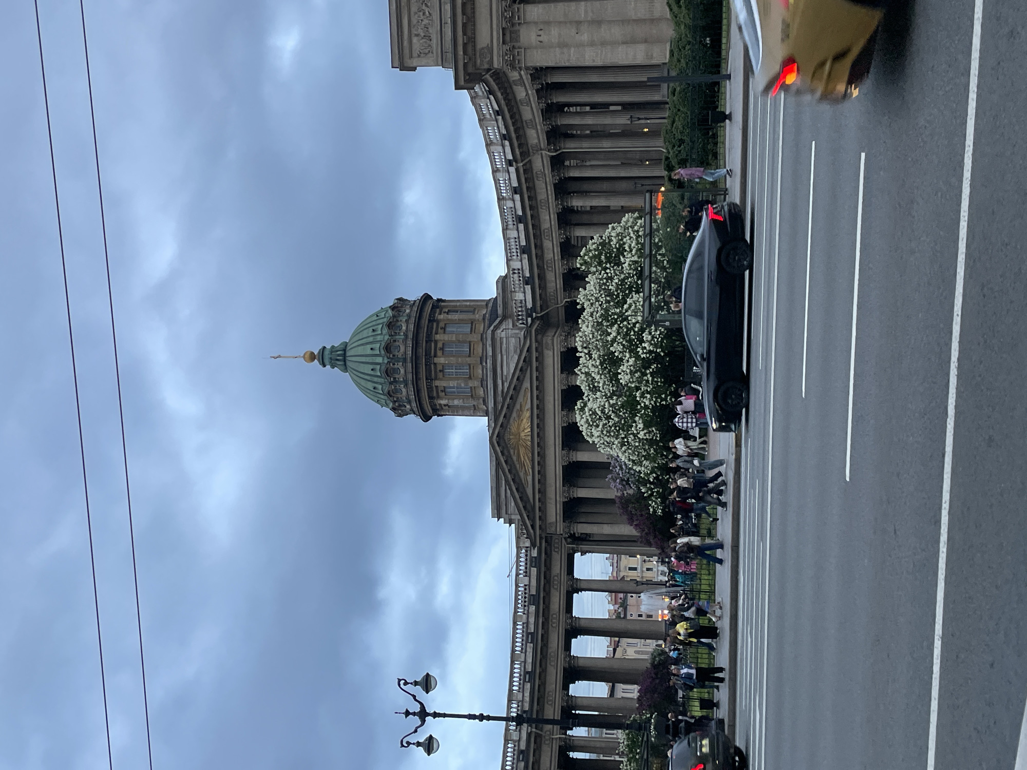 The image captures a view of the Kazan Cathedral in Saint Petersburg, Russia. The cathedral, with its iconic dome, is visible against a cloudy sky. The camera is at street level, showing a road with lane markings. Several people are walking along the side of the road and in front of the cathedral. Cars are present on the road, including a black car stopped at the side and another car driving. Trees are in full bloom. A street lamp can be seen in the foreground. Electrical wires run across the left of the image.