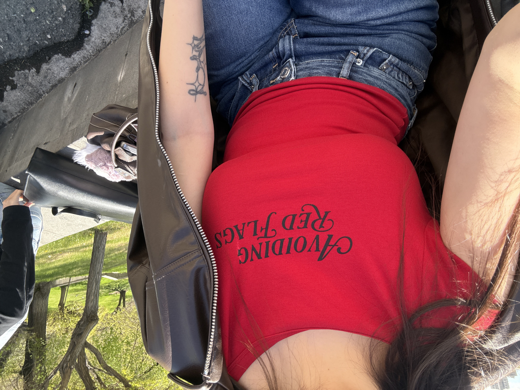 The image, corrected from its 90-degree clockwise rotation, captures a person relaxing outdoors in Yerevan, Armenia, on a bright, sunny day. The subject, visible from the chest up, wears a vibrant red, ribbed crop top emblazoned with the black script text "AVOIDING Red FLAGS." They are dressed in blue denim jeans and a dark brown or black leather-like jacket with a prominent silver zipper running down the front.
Long, dark hair spills over one shoulder and arm. A distinct, intricate tattoo, featuring an abstract, possibly ornamental or spiderweb-like design, is visible on their left forearm. Nearby, two handbags are present: a dark, structured one and a smaller brown or bronze purse with a light-colored, fluffy trim.
The background reveals a concrete sidewalk or pathway adjacent to a lush green area dotted with trees, some showing fresh spring foliage. The bright sunlight creates clear shadows, indicating fair weather. The overall scene suggests a casual and relaxed moment in an urban park or public outdoor space.