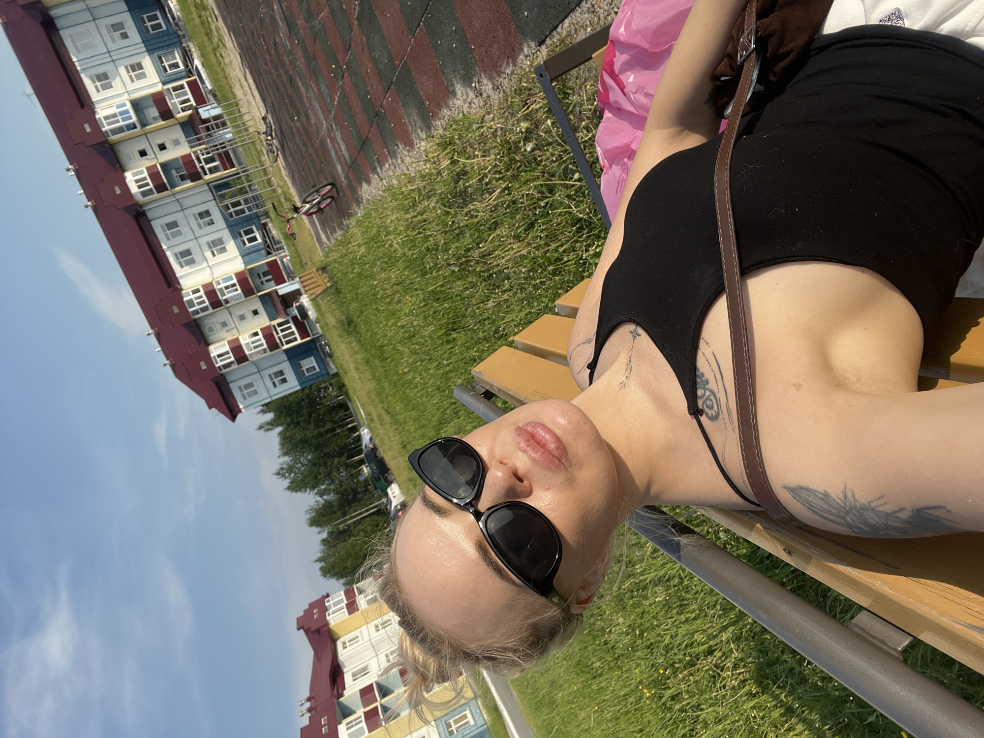 In an outdoor urban setting under a bright sky, a young woman with blonde hair pulled up in a bun is seated on a park bench. She is wearing large black sunglasses and a black tank top, and has several tattoos visible on her arm and chest. A brown strap from a bag is visible across her chest. The bench is wooden and weathered. Behind her, a playground area with red and black rubberized surfacing is visible, with a bicycle lying on its side. Further in the background are apartment buildings with colorful facades and red roofs, surrounded by green grass and trees. The environment appears to be a residential area with a playground.