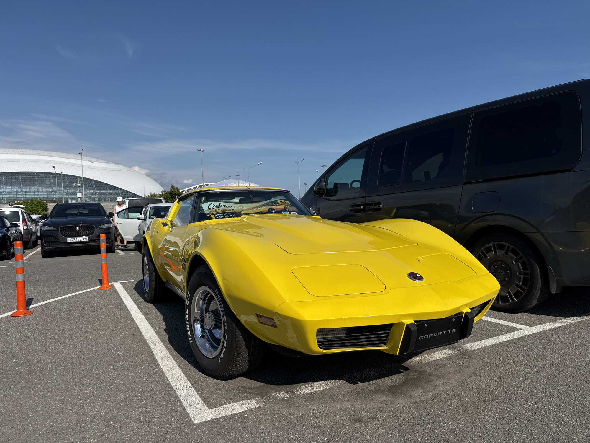 The image displays a bright yellow Chevrolet Corvette C3, prominently parked in an asphalt lot under a clear blue sky with sparse clouds. The location is Sirius, Russia, identifiable by the distinctive white-domed Fisht Olympic Stadium visible in the background.
The Corvette features "CORVETTE" on its front license plate mount and "COOPER G/T RADIAL" inscribed on its tire sidewalls. A "Cabrio 123" sticker, containing Russian text and phone numbers (specifically 8 (988) 402-00-00 and 8 (988) 120-00-00), is affixed to its windshield, along with a smaller yellow sticker indicating "50+".
To the right of the Corvette, a dark-colored minivan or large SUV is partially visible. Behind the Corvette, a black Jaguar SUV, bearing Russian license plate H372CE 193, is parked. Further in the background, a male individual wearing a white cap and a white shirt stands between the black Jaguar and a white minivan, interacting with an open car door.
Two orange traffic bollards are positioned near the black Jaguar. The parking lot surface is asphalt, marked with white painted lines delineating parking spaces. Several tall streetlights are visible in the distance. The scene is brightly illuminated by sunlight, casting distinct shadows.