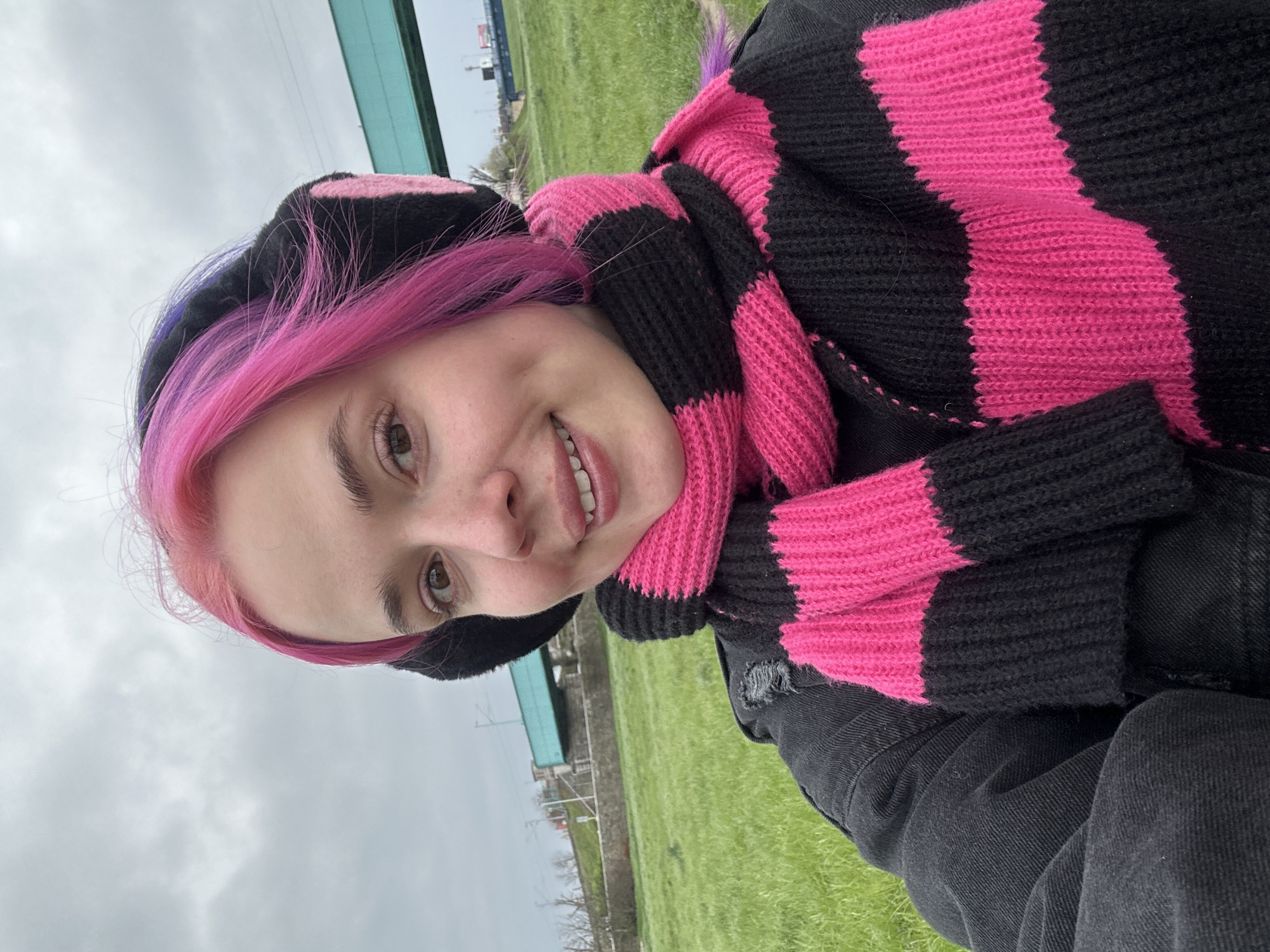A young person with pink hair stands on a grassy area with buildings in the background. The person wears a black and pink striped scarf and a black hat with pink accents. They are smiling. The location appears to be outdoors, likely in a park or open space within a city. Given the instructions, the city is Zagreb, Croatia. The sky is overcast. The person is also wearing a black jacket.