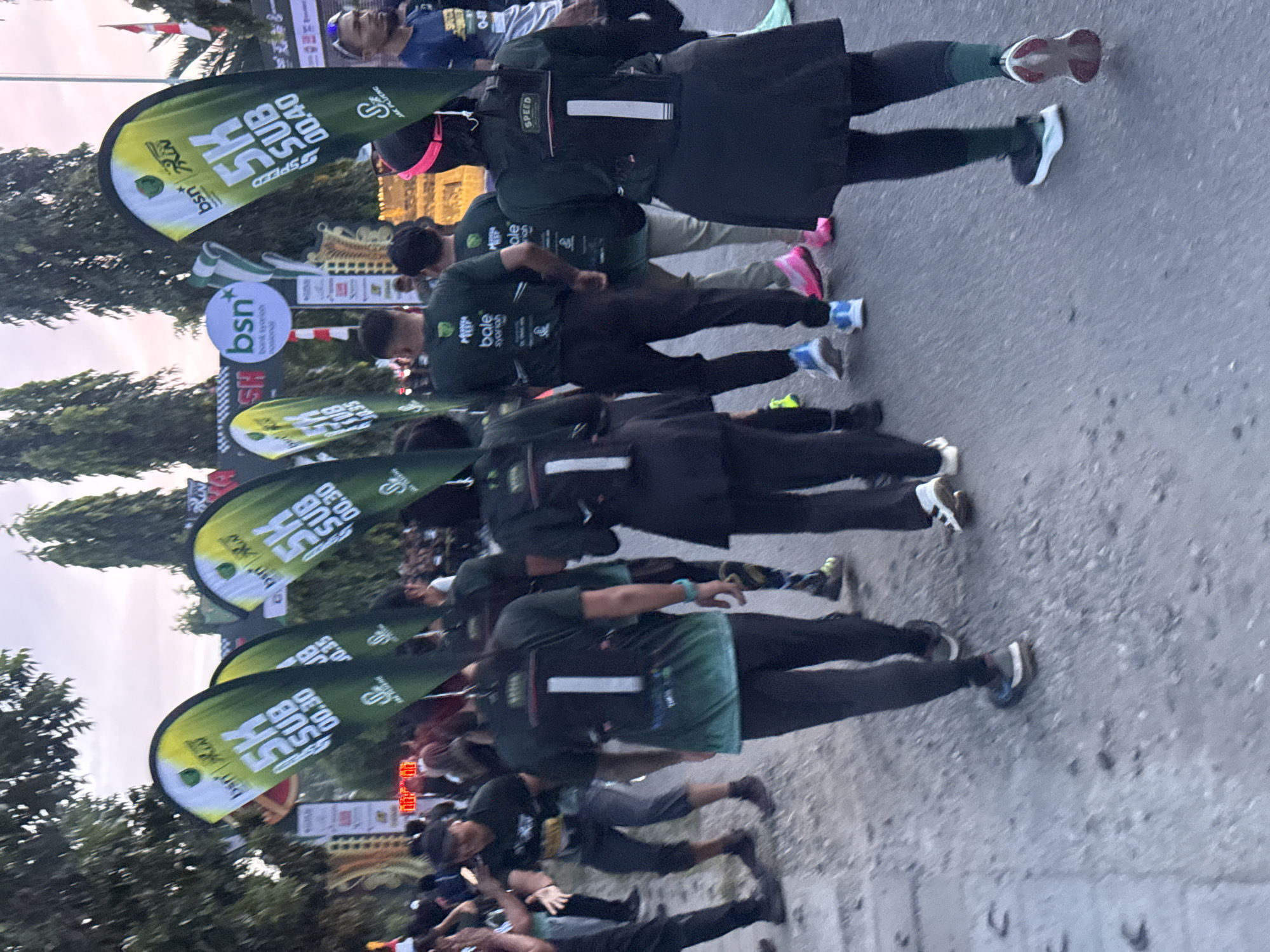 The image captures a group of people participating in a 5K run event, likely held outdoors in the city of Lhokseumawe, Indonesia, given the Indonesian language on the banners. The scene appears to be at the start or a checkpoint of the race, with several participants in the foreground, mostly facing away from the camera.
In the foreground, several individuals are walking or standing, some wearing dark green athletic attire with white numbers on the back, indicating they are runners. The numbers visible are "1" and possibly others obscured. One runner in the center-right is wearing a black jacket with "SPEED" written on the back in white. Another runner on the far right is wearing a blue shirt and sunglasses. The participants are on a paved road, with trees and other foliage visible in the background.
Dominating the upper portion of the image are several large, vertical banners. These banners are green and yellow, displaying "BSN", "5K", "SUB 00.30", and "SUB 00.40" in white text. The "BSN" likely refers to Bank Syariah Nasional, a common sponsor of such events in Indonesia. The banners suggest a timed race category.
In the middle ground, there are more people, some appearing to be spectators or event organizers. Further back, there is a structure with checkered patterns and what appears to be a finish line or an event entrance arch. The sky is overcast, suggesting it could be early morning or late afternoon. The overall atmosphere seems to be one of an organized sporting event. The ground is paved and appears slightly dusty.