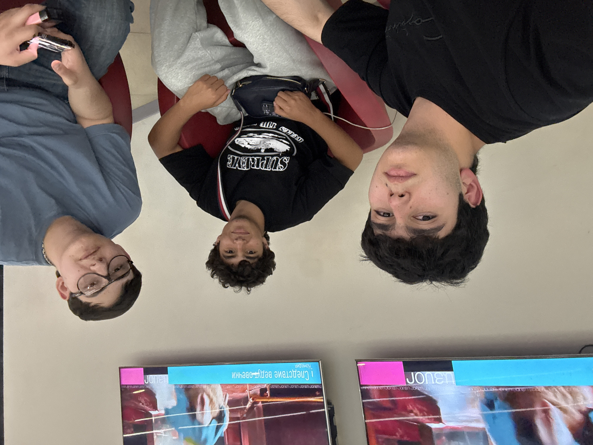 Three young males are sitting in what appears to be a waiting area in Yerevan, Armenia. They are positioned in a row of red chairs. The individual on the left wears glasses and a blue shirt, holding a small electronic device. The central figure is dressed in a black Supreme t-shirt and grey sweatpants. The third person is wearing a black t-shirt. In front of them, two televisions display content in what appears to be a public space. The wall is a light cream color. The inverted text on the TV screen indicates the photograph was taken upside down.