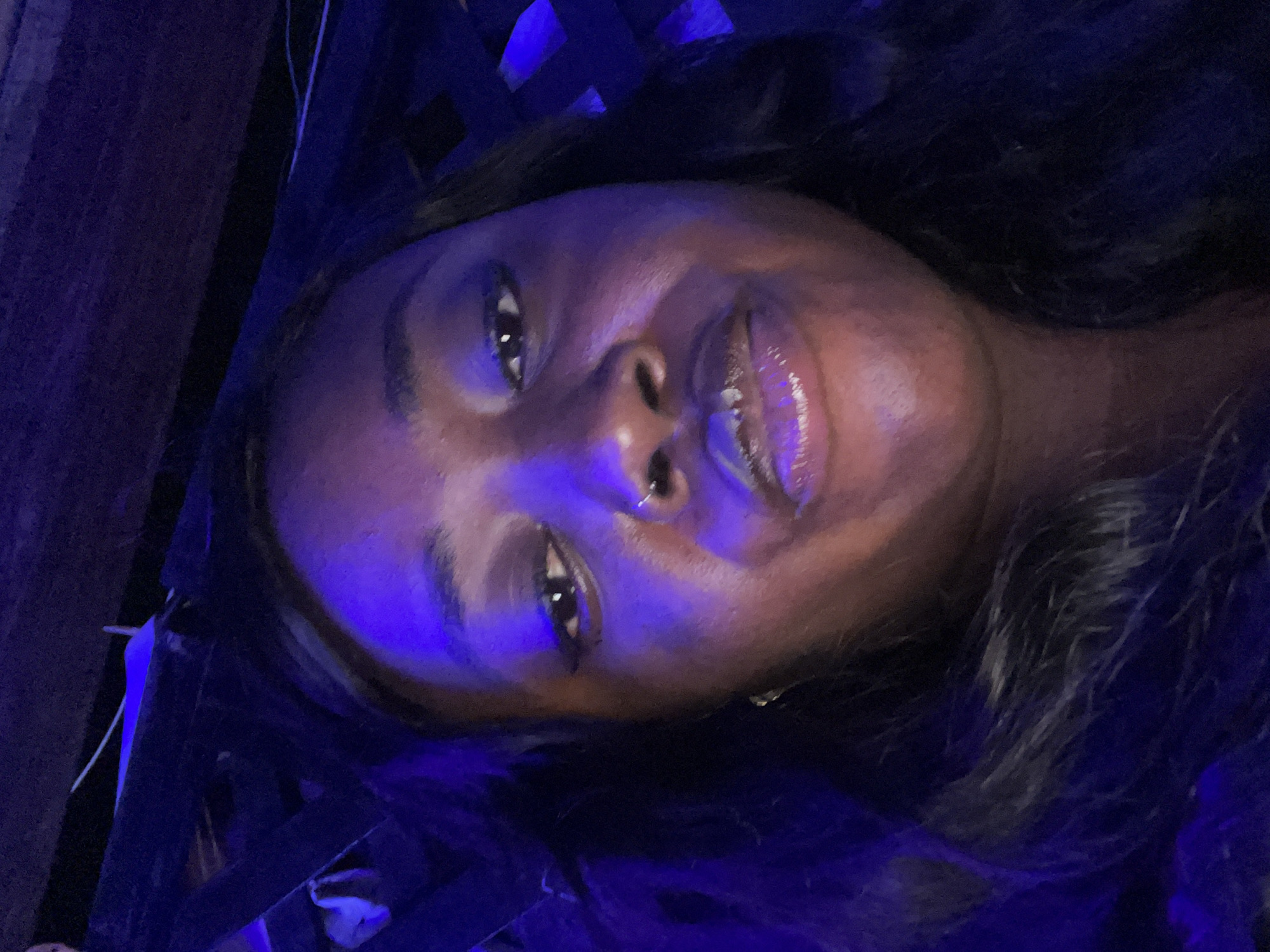 A close-up selfie shows an adult individual, appearing female, directly facing the camera with a subtle, closed-mouth smile. The subject has dark skin and dark, wavy hair cascading down both sides of her face, with portions illuminated by the ambient light. Her eyes are dark, with visible eyeliner, and she wears a small silver hoop nose ring in her right nostril. Her lips are full and appear to have a glossy, possibly shimmering, product applied. A small gold-colored hoop earring is visible on her right earlobe. The scene is dominated by strong artificial blue/purple lighting, which casts a distinct hue across the subject's face and creates deep shadows in the background. The environment behind the subject is dark and indistinct, revealing shadowed architectural elements. These include a dark, slatted or lattice-like structure in the upper right portion of the frame, and a less defined dark surface with a faint white line in the upper left, suggesting an indoor or semi-enclosed commercial venue like a lounge or club in the city of Abuja, Nigeria.