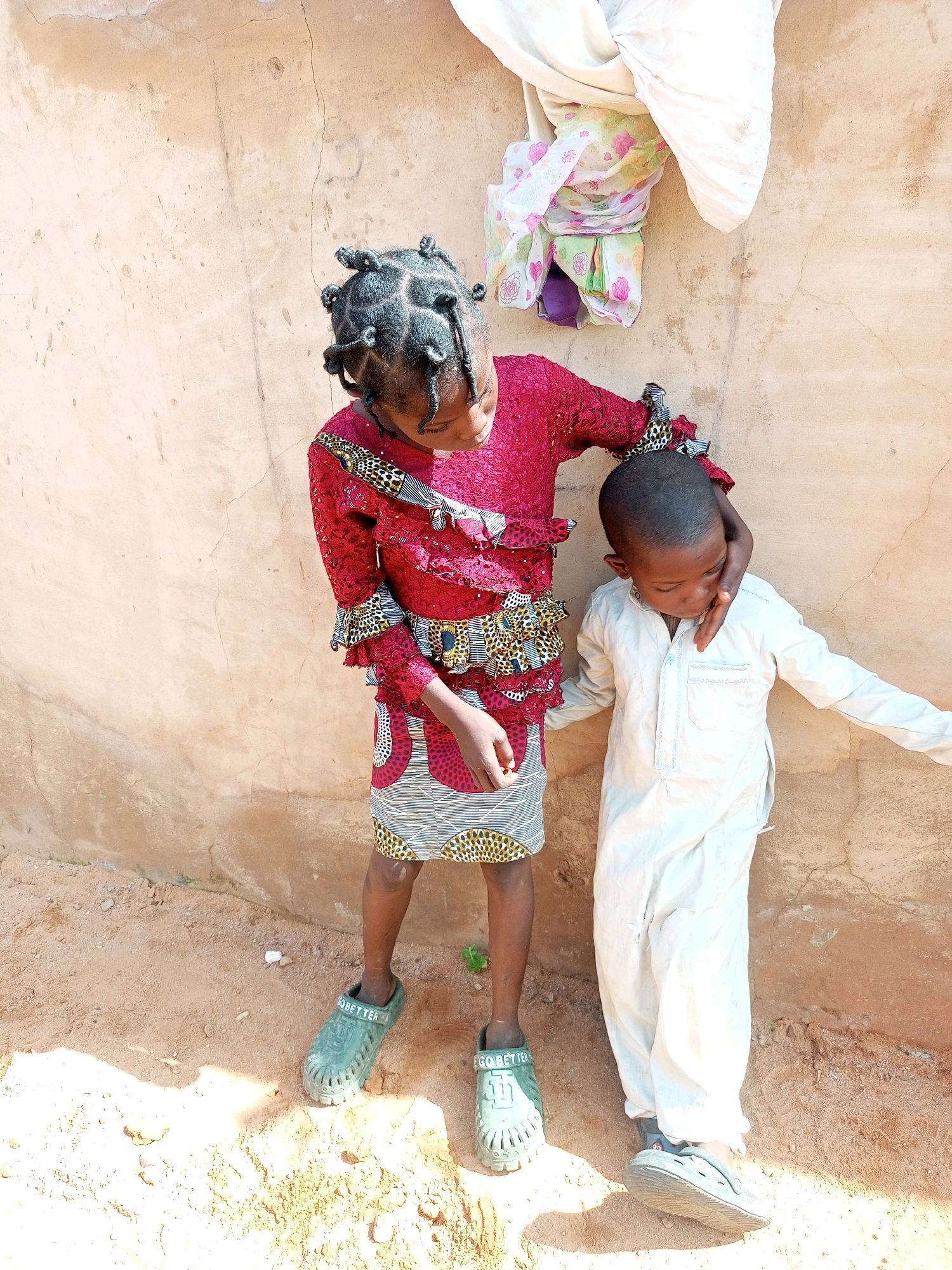 In Gombe, Nigeria, two children stand against a textured, light brown wall. The child on the left, a girl, wears a vibrant red, ruffled top with a patterned skirt. Her hair is styled in numerous small, dark braids. She has her left arm around the neck of the child on the right, a boy. The boy is dressed in a light-colored, traditional garment and his head is bowed. His right arm is extended to the side, and his left arm is resting on his lap. The ground beneath them is composed of sandy soil with some small pebbles and debris. Above them, on the wall, a bundle of white and patterned fabric is draped. The overall scene suggests an outdoor setting in a built environment.