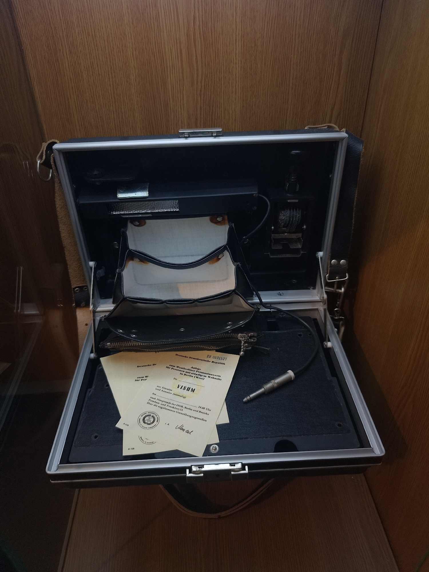 An open briefcase with a dark gray interior and a silver trim is positioned against a wooden surface. Inside the briefcase, a black pouch with white accents and a zipper is visible. A silver audio jack connector is lying on the right side. Several sheets of paper with German text and official stamps are placed on the bottom-left side of the briefcase. The paper is a "VISUM" document from the Deutsche Demokratische Republik for entry and exit. The setting is likely indoors, possibly within a museum or personal collection.
