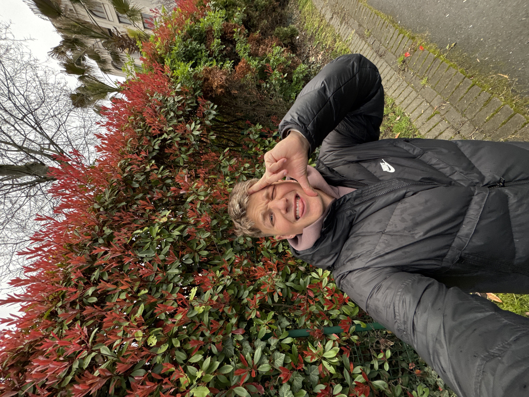 The image shows a young person taking a selfie outdoors next to a hedge with red and green leaves. The person is wearing a black puffer jacket with a Nike logo and is making a playful face and a "peace" sign with their hand. The background includes a sidewalk, some buildings, and other greenery. Although the location is stated as Burgas, Bulgaria, this cannot be definitively confirmed from the image alone. Further features are a dry tree in the background and a building on top of the hedge. The sidewalk looks weathered and there is moss on it.