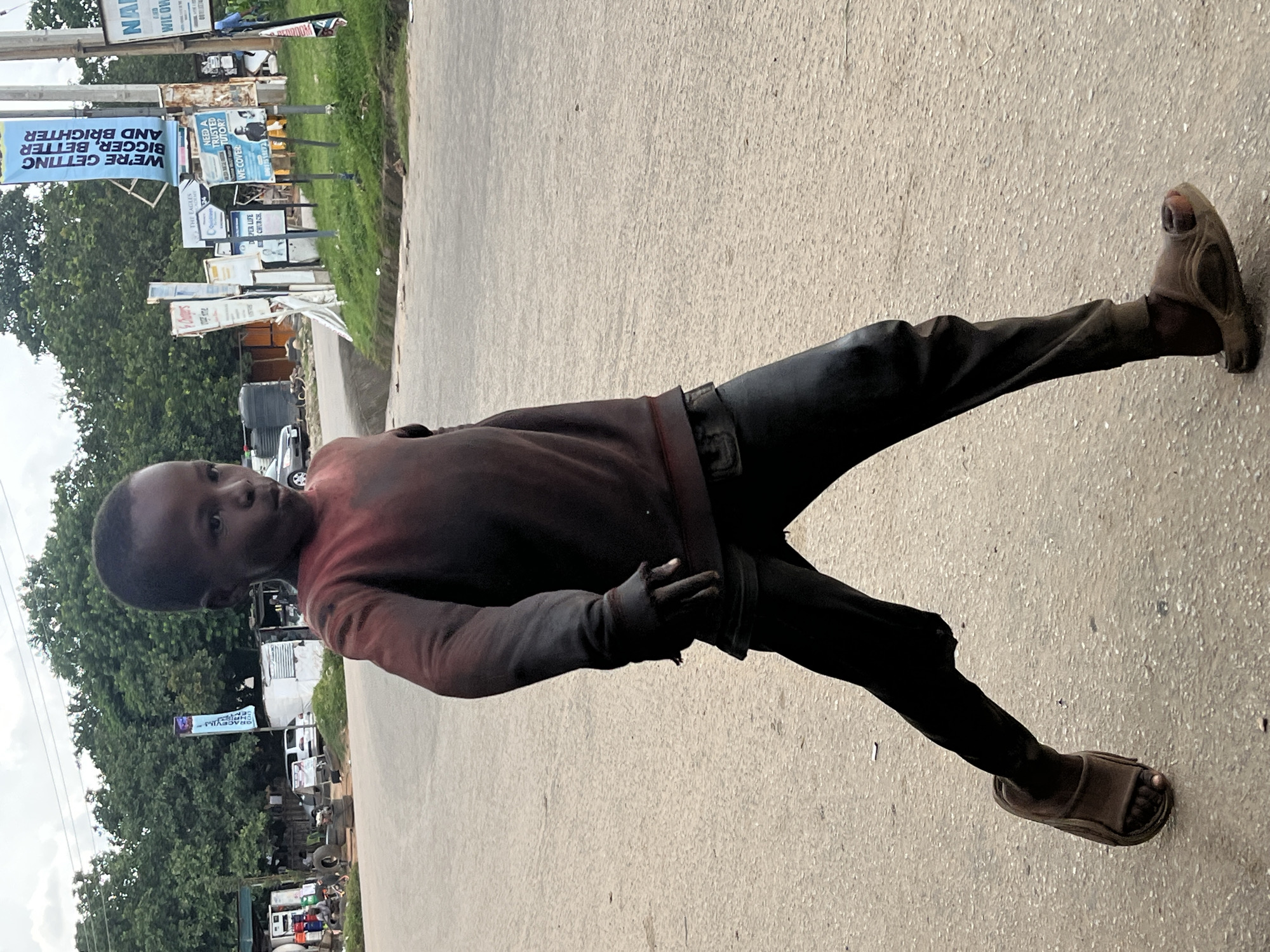 In Abuja, Nigeria, a young boy stands on a paved road, his body angled towards the camera. He wears a dark red long-sleeved shirt and dark pants, with brown sandals on his feet. The boy looks directly at the viewer with a neutral expression. The scene appears to be outdoors during the daytime, with overcast skies suggesting a possibly humid environment.
Behind the boy, a row of billboards and signs are visible. One prominent blue banner reads, "WE'RE GETTING BIGGER, BETTER AND BRIGHTER". Other signs, partially obscured, appear to advertise services like tutoring, with one specifically asking, "NEED A TRUSTED TUTOR?". The setting seems to be a roadside area with some vegetation and trees, indicating a less urbanized or commercial part of the city. Several vehicles, including a white van and what appears to be a black car, are parked in the background. The overall impression is of a busy roadside environment with various advertisements.