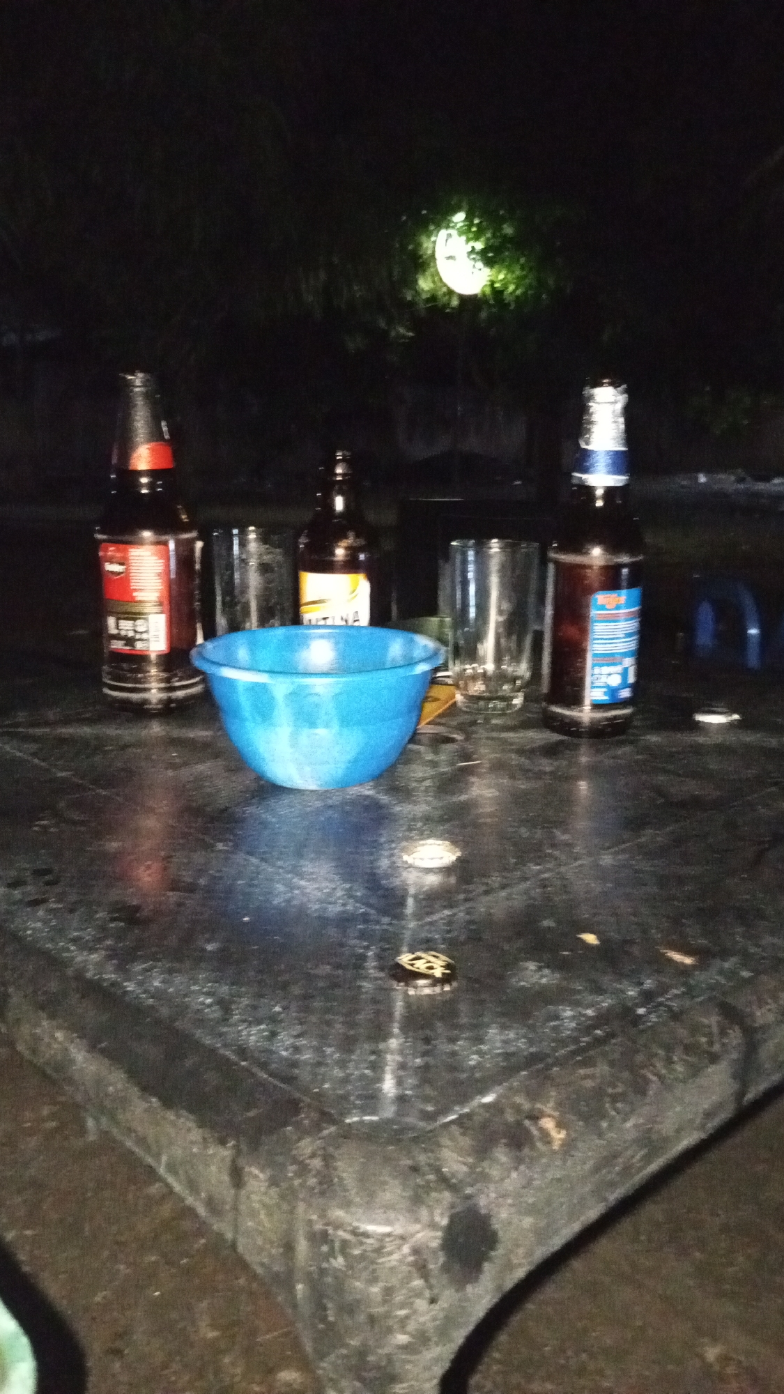 This image, captured at night in Adankolo, Nigeria, depicts an outdoor scene centered on a dark, possibly plastic, rectangular table. On the table are three upright glass bottles: one with a red label (identifiable as Star beer), one with a yellow label (identifiable as KUNNA), and one with a blue and white label. Two empty, clear glass tumblers are also present alongside a central blue plastic bowl. Several metallic bottle caps are scattered across the table's surface. The background is dark, featuring indistinct foliage illuminated by a single, bright, circular light source. No individuals are present in the frame. The table surface appears somewhat wet or stained.