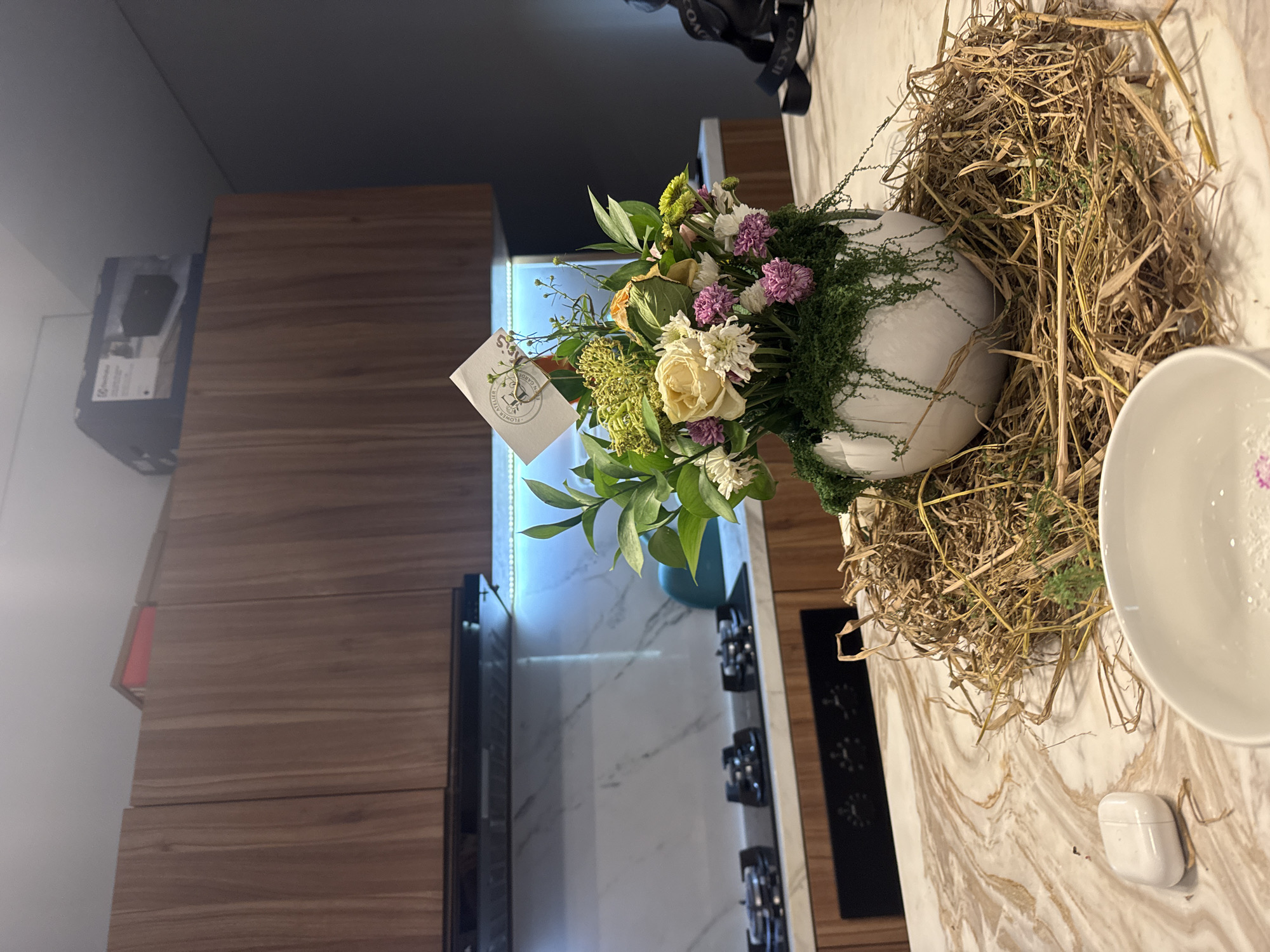 The image shows a bouquet of flowers in a white, round vase. The flowers are a mix of white, yellow, and purple, with green leaves. The vase sits inside a nest of dried grasses. A small white card with the "FLOWER ATELIER" logo is attached to the bouquet. To the right is a white bowl and a pair of white earbud case. The surface is marble or a similar material. In the background is a kitchen with wooden cabinets and a stovetop, with a marble backsplash. The location appears to be Kerobokan, Indonesia based on the "FLOWER ATELIER" card, indicating a potential local business.