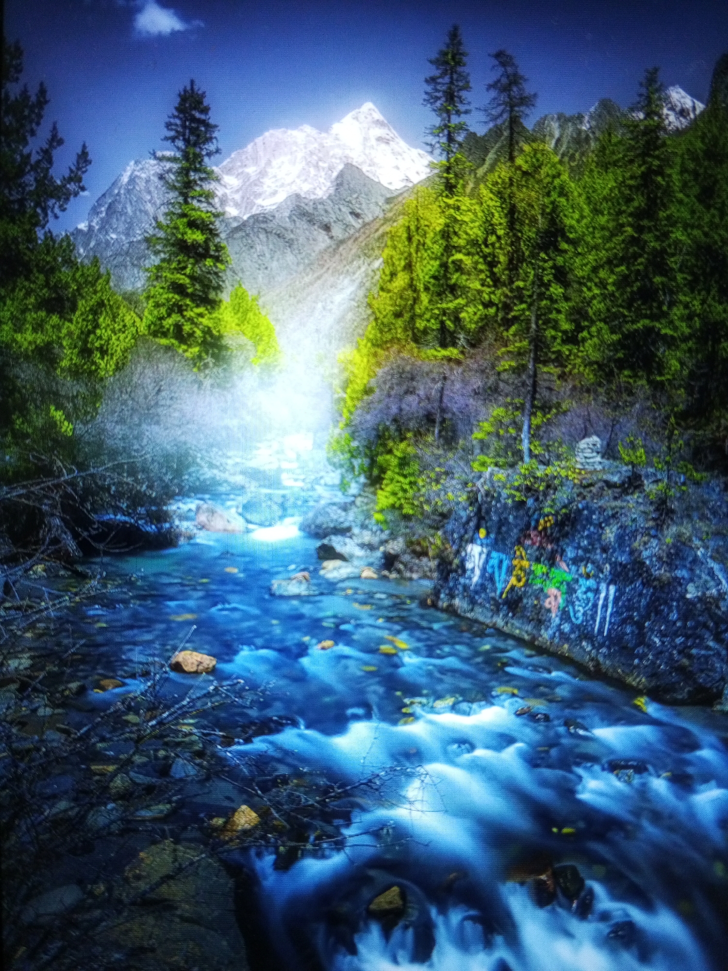 This media file captures a vibrant mountainous landscape with a prominent river flowing through a lush valley, depicting a scene possibly located in Tinaquillo, Venezuela.
The central subject is a fast-flowing river, rendered with a long exposure technique that creates a smooth, misty, and ethereal appearance for the water, predominantly in shades of blue and white, with visible rocks and submerged branches. The river originates from a bright, glowing mist in the mid-ground, suggesting a powerful light source or atmospheric effect.
In the background, imposing snow-capped mountains rise majestically against a deep blue sky, which features a single white cloud in the upper left corner. The slopes and riverbanks are densely covered with vibrant green foliage, including tall evergreen trees and bushy undergrowth.
A notable detail on the right bank of the river is a dark rock face adorned with colorful graffiti or symbolic markings. These include abstract designs and possibly stylized figures in bright yellow, green, blue, and white. Various rocks and stones are scattered within the river and along its edges.
The image conveys a sense of serene natural beauty and grandeur. The time of day appears to be daylight, with clear weather conditions allowing for a vivid display of colors and atmospheric light. No discernible text is visible, only the colorful symbols on the rock.