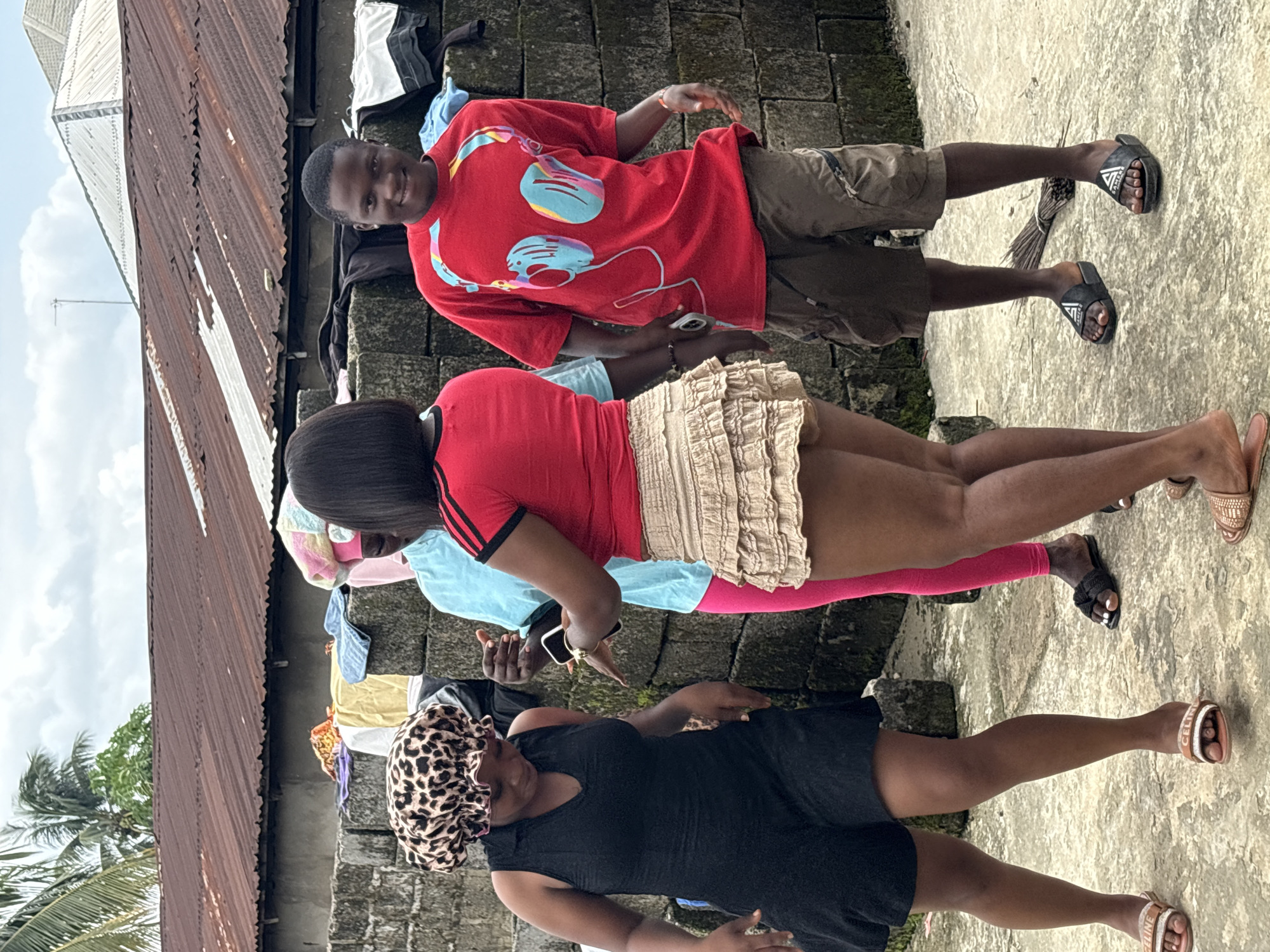 This daylight outdoor photograph, taken in Ikot Ekpene, Nigeria, features four individuals – one man and three women – standing on a textured concrete or stone ground.
In the back-right, a man with short black hair is smiling and looking towards the camera. He wears a red short-sleeved t-shirt with a colorful graphic depicting headphones in blue, pink, and yellow. He also wears khaki shorts and black slide sandals with a white triangular logo that might read "SAVED."
To his left and slightly in front, a woman stands dressed in a red short-sleeved top with black stripes on the sleeves, a ruffled beige mini-skirt, and pink leggings or trousers. She holds a white smartphone with gold trim in her hands, looking downwards. She wears black slide sandals with a white detail.
Closest to the camera, a second woman wears a black sleeveless dress and a leopard print head covering. She looks downwards or slightly away from the camera. Her feet are in light brown flat sandals with a white strap, potentially bearing the text "CELINE." Partially visible behind the central woman is a third woman, wearing a light blue top and what appears to be pink leggings.
The background on the left side features a rusted corrugated metal roof, sloping downwards, against a bright sky with white clouds. A palm tree and a small antenna are also visible. On the right, a gray cinder block wall is topped with an assortment of folded clothes in various colors including white, blue, yellow, and pink, suggesting a casual outdoor setting or drying area. The bright lighting indicates it is daytime.