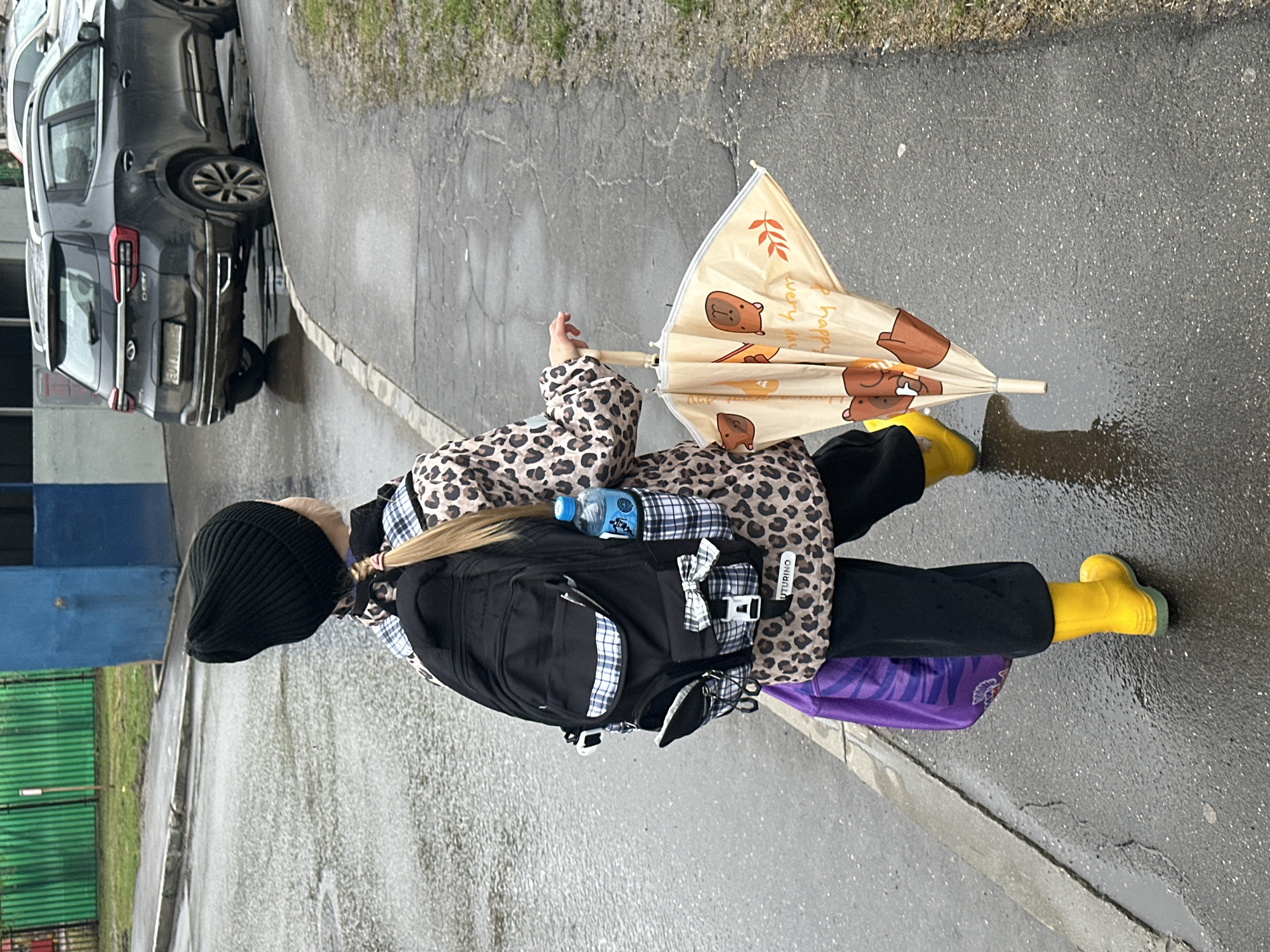 A girl is walking on a wet, urban sidewalk. She is wearing a black beanie, a leopard-print jacket, black pants, and bright yellow rain boots. She has a black backpack with a water bottle attached and is carrying a closed umbrella with cartoon bears printed on it. She is also carrying a purple bag. Cars are parked nearby, and there is grass and a blue building with green fencing visible in the background. The photograph was taken on the street in Moscow, Russia.