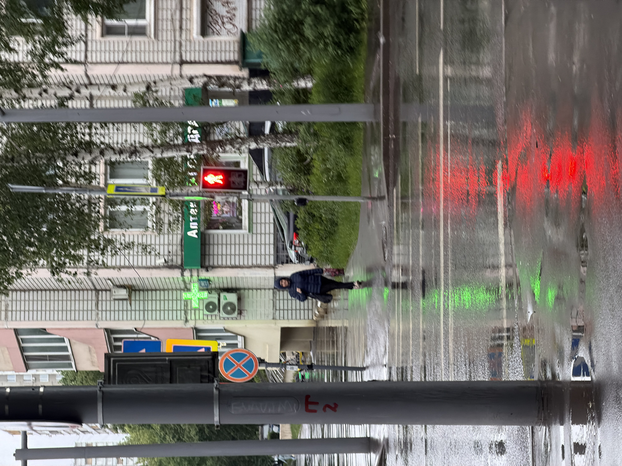 The image shows a street scene in Moscow, Russia on a rainy day. The view is from behind a gray pole, which has red graffiti on it. A pedestrian is crossing the street. A traffic light displays a red "walk" signal. The street is wet and reflects the traffic lights as blurred green and red lines. Buildings line the street, including a pharmacy ("Аптека") with a green cross sign. A "no stopping" sign is visible.